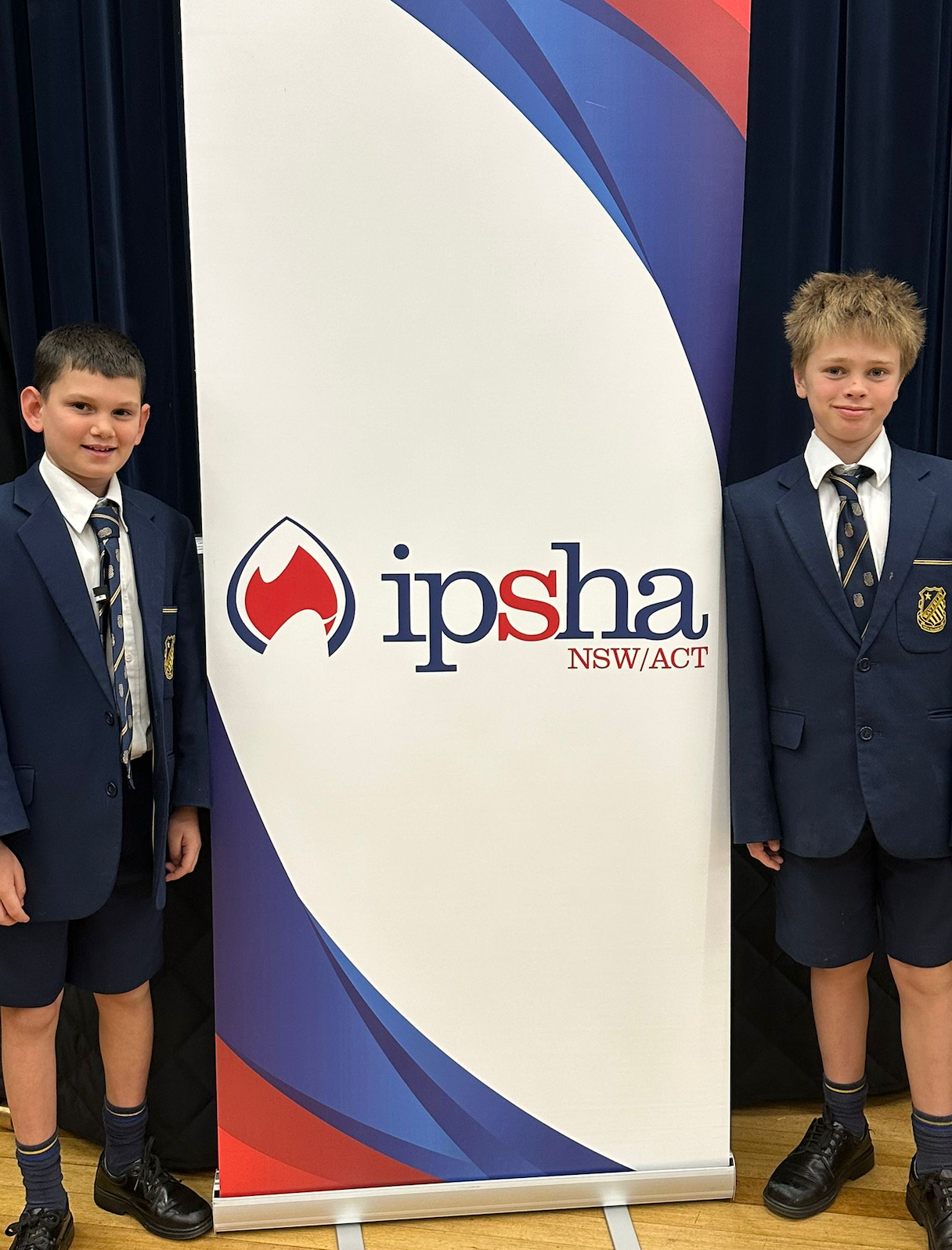 Two boys in navy blue school uniforms stand on either side of a banner that reads “ipsha NSW/ACT.” The banner has a blue, red, and white design. They are indoors with a dark curtain behind them.
