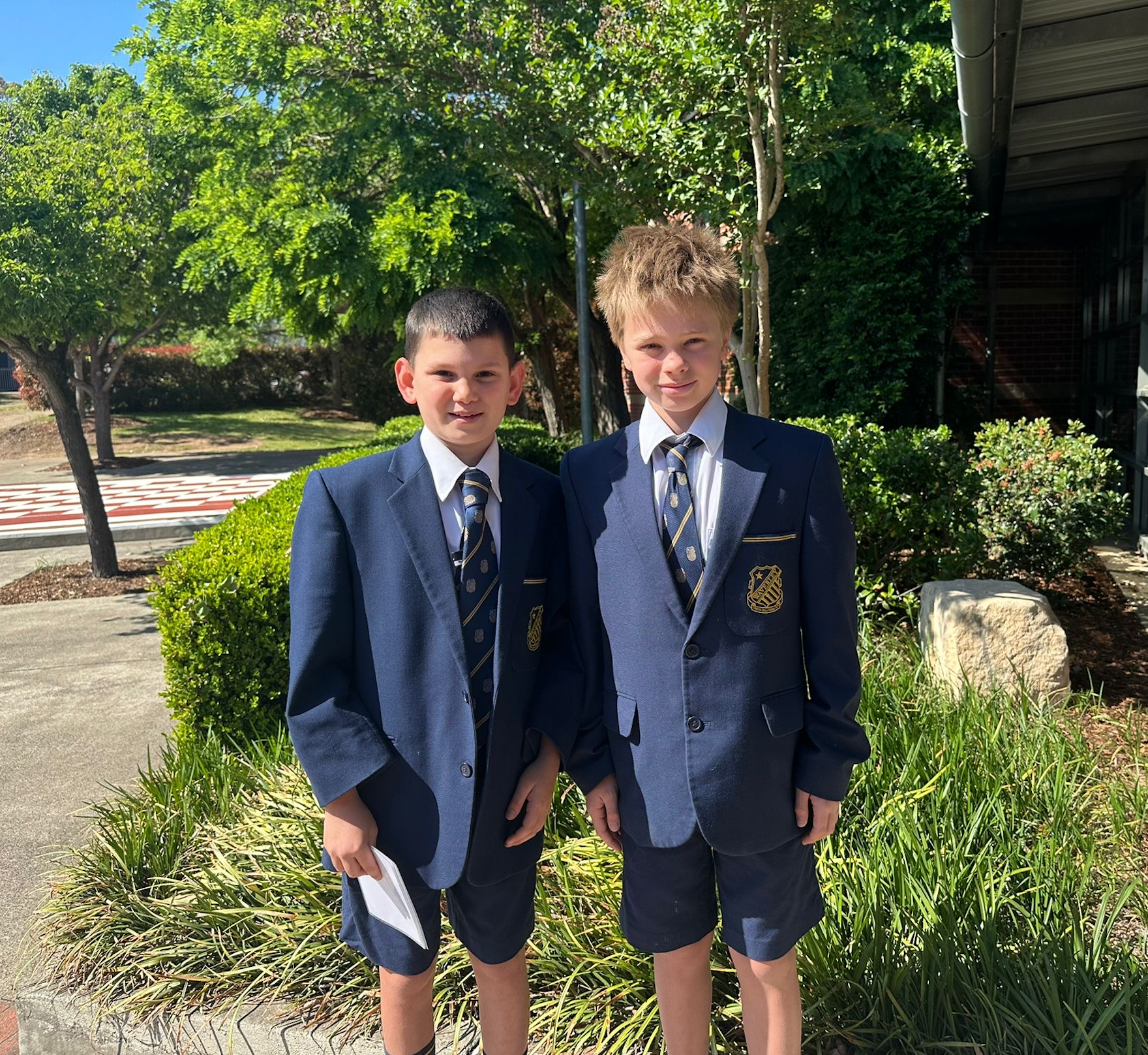Two boys wearing blue school blazers, ties, and shorts stand side by side outdoors, smiling. They are in front of bushes and trees on a sunny day, with a building partially visible to the right.
