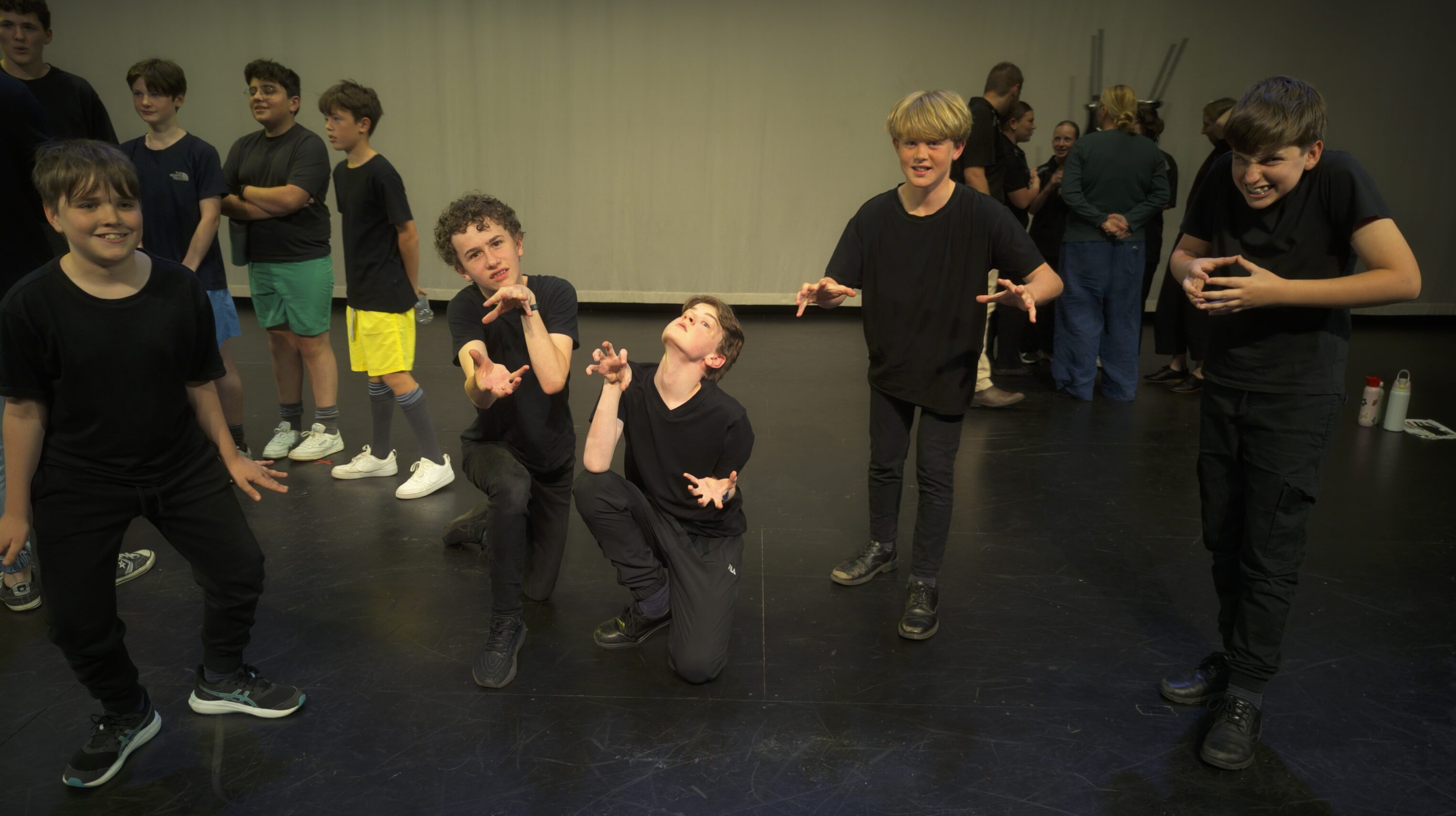 A group of boys in black clothing pose animatedly on a stage, with several others talking in the background. The boys in front make expressive gestures and facial expressions, as if performing or acting out a scene.