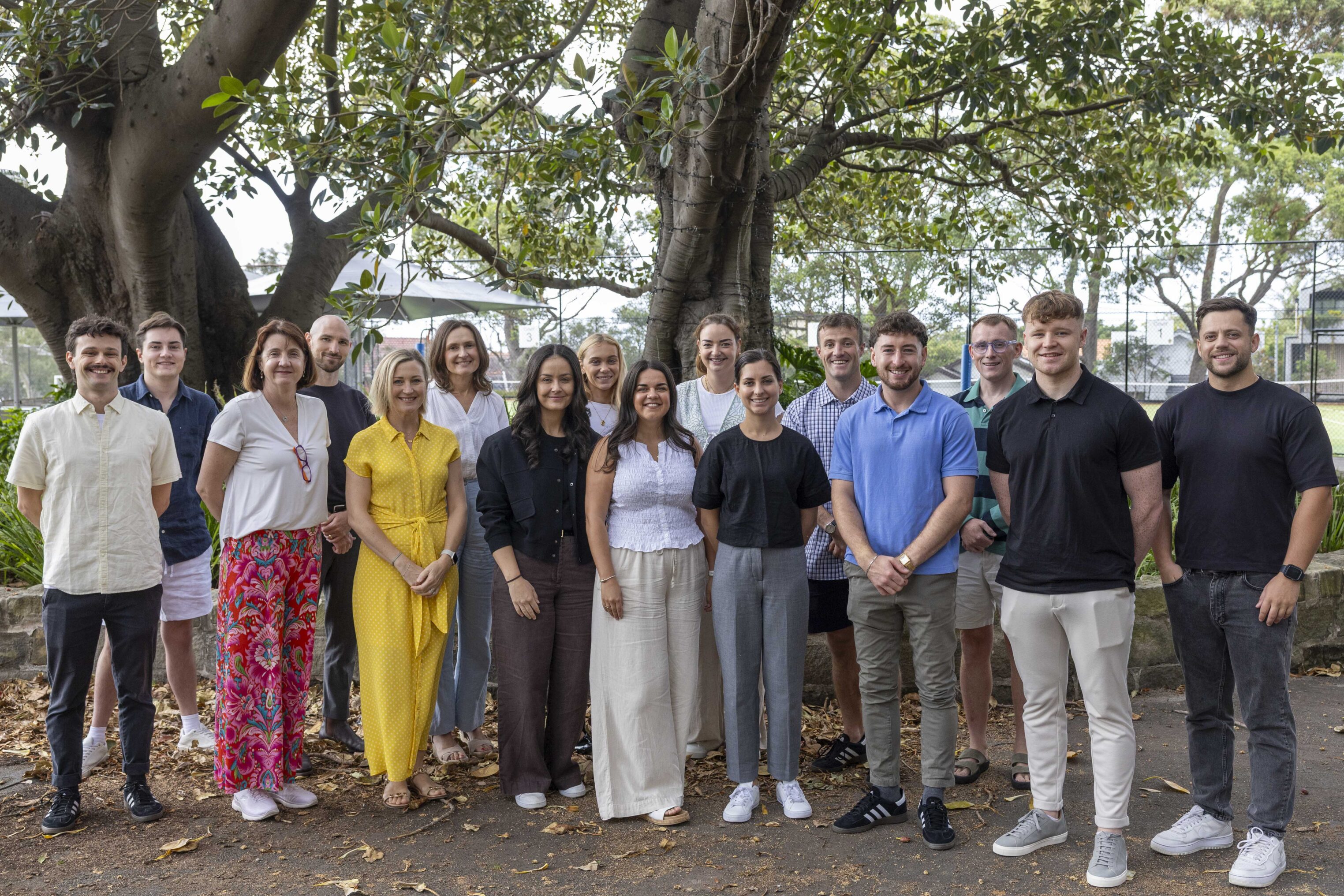 A group of sixteen people, a mix of men and women, stand outdoors in front of a large tree, smiling at the camera. They are dressed in casual, summery clothes, and the background includes greenery and park features.
