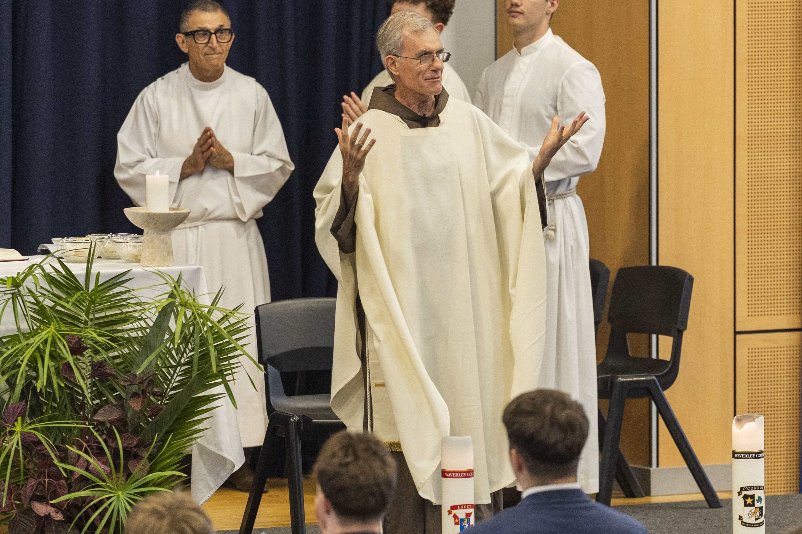 A priest in cream robes stands with arms raised, addressing seated people during a religious ceremony. Two other clergy in white robes stand behind him near a table with a lit candle and greenery.
