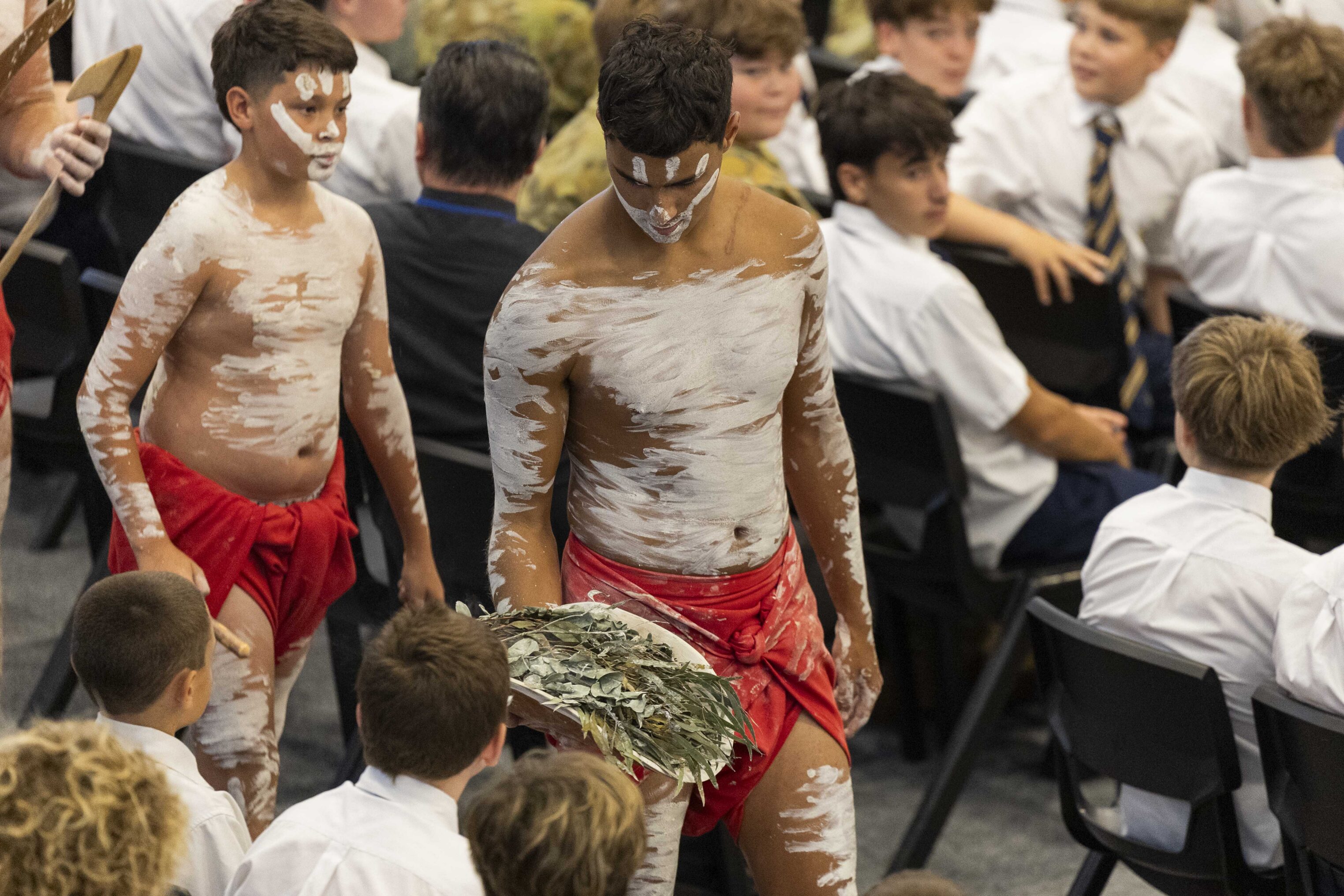 Two young men wearing red cloths and traditional white body paint walk through a seated crowd of schoolboys in white shirts; one carries a bundle of green leaves, possibly for a ceremony or cultural event.