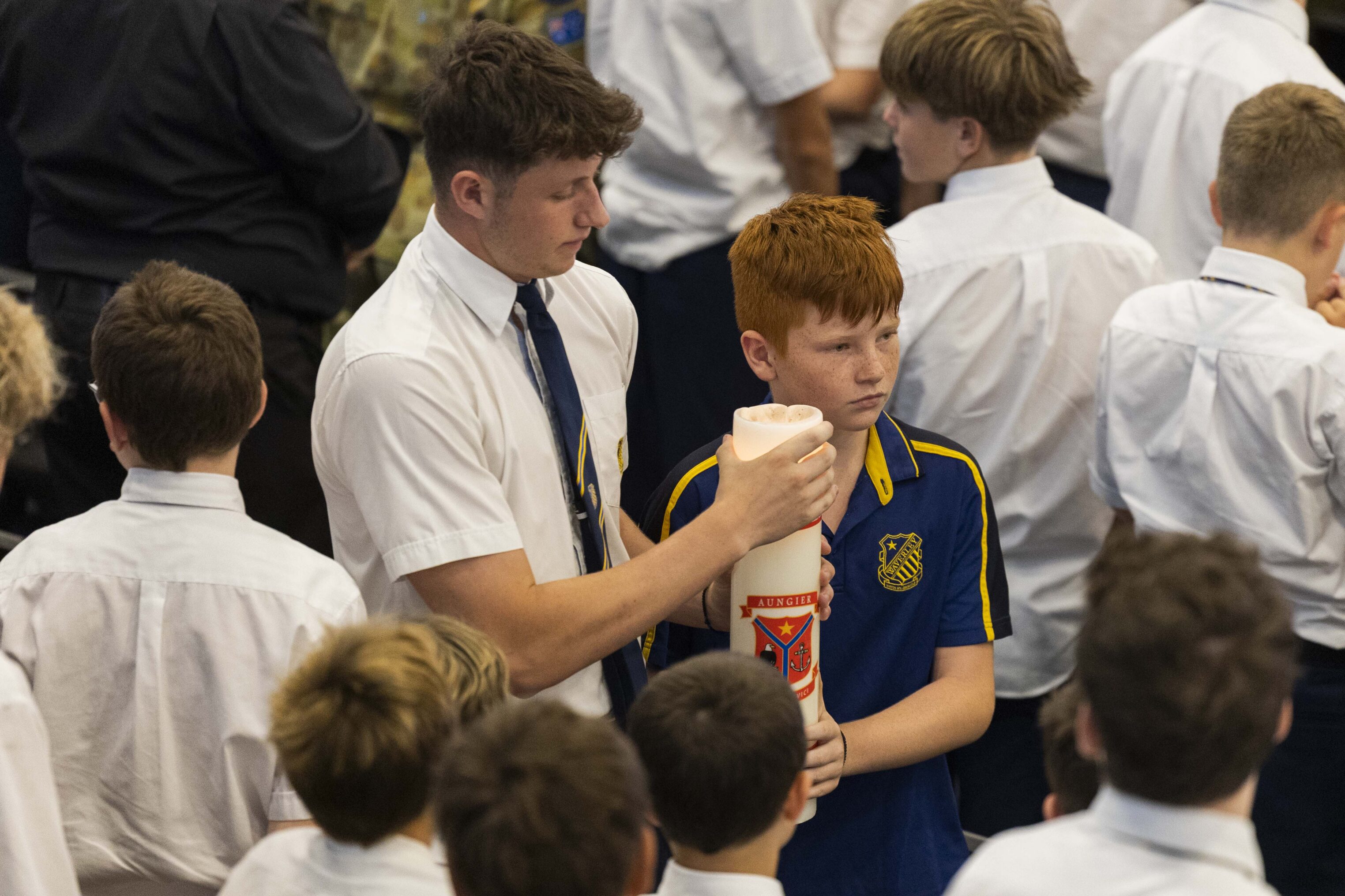 A group of boys stands together; one boy in a white shirt and tie helps a boy in a blue sports shirt hold a large white candle. The scene appears to be at a school event or ceremony.
