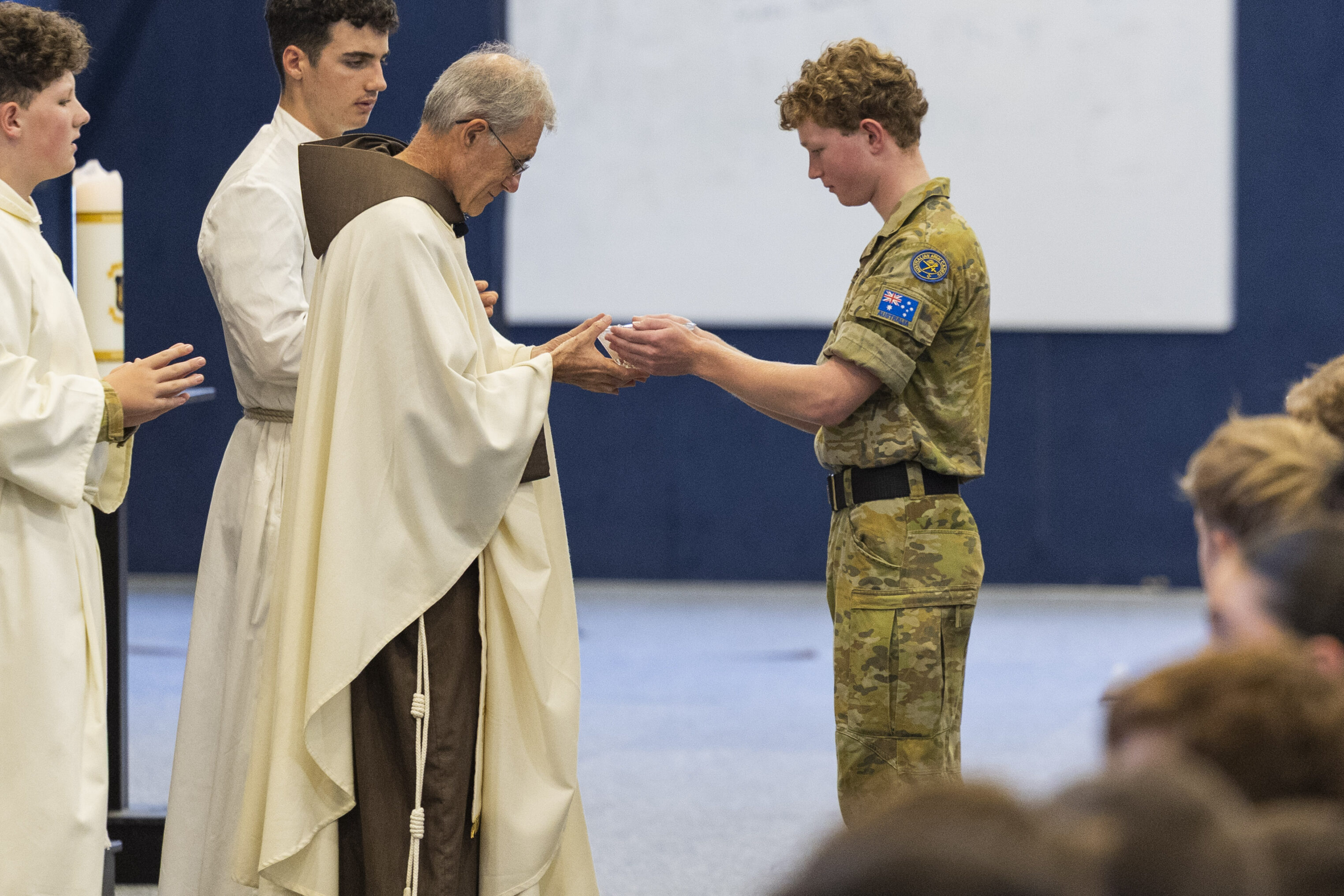 A soldier in camouflage uniform receives communion from a priest dressed in brown and white robes during a ceremony, with other people in white robes standing nearby.