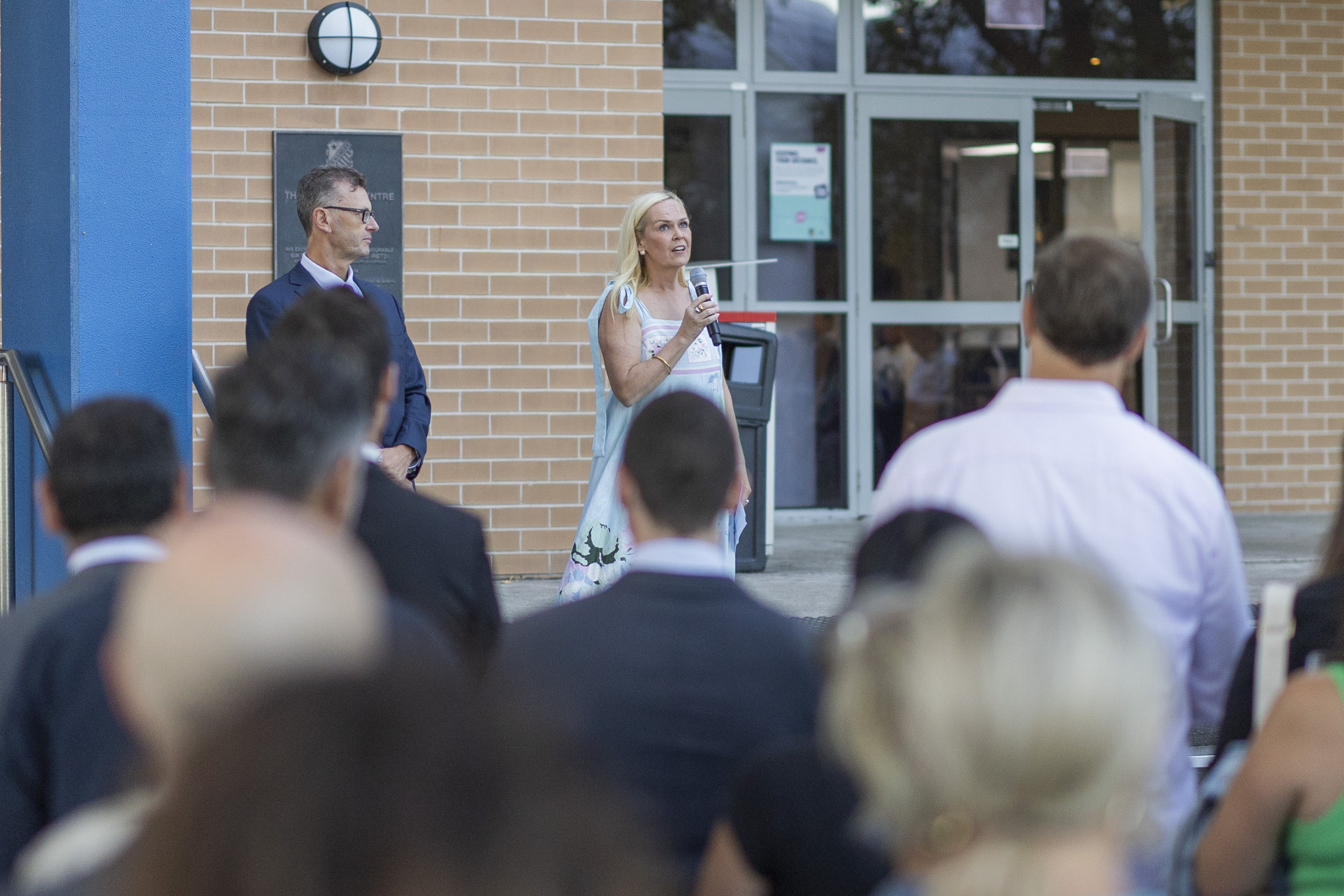 A woman speaks into a microphone to a crowd outside a brick building, whilst a man stands beside her. The audience stands facing them, and the scene appears to be an outdoor event or gathering.