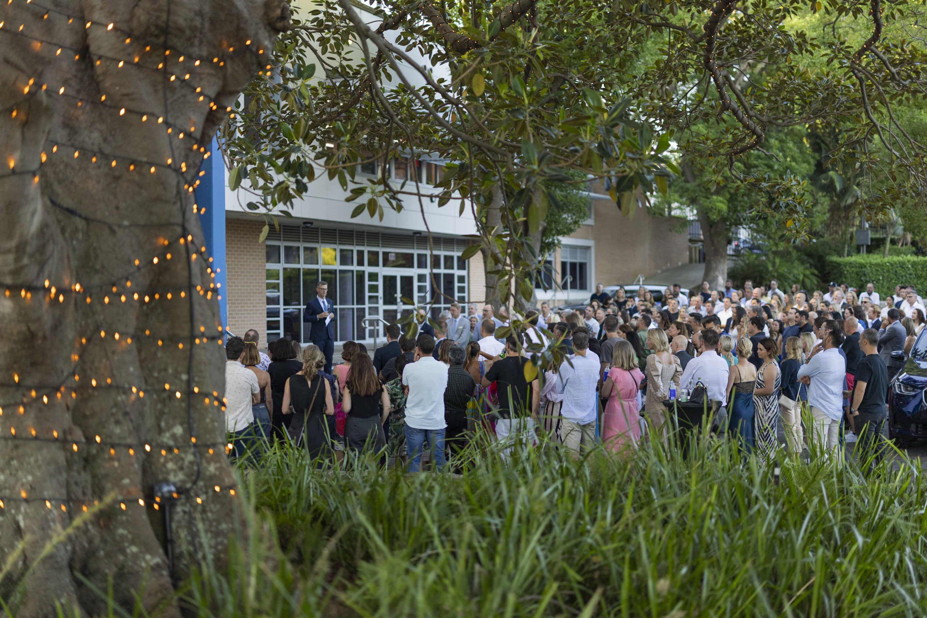A large crowd gathers outdoors near a building, listening to a speaker in a suit. Fairy lights wrap around a tree in the foreground, and greenery surrounds the scene. The atmosphere appears festive and sociable.