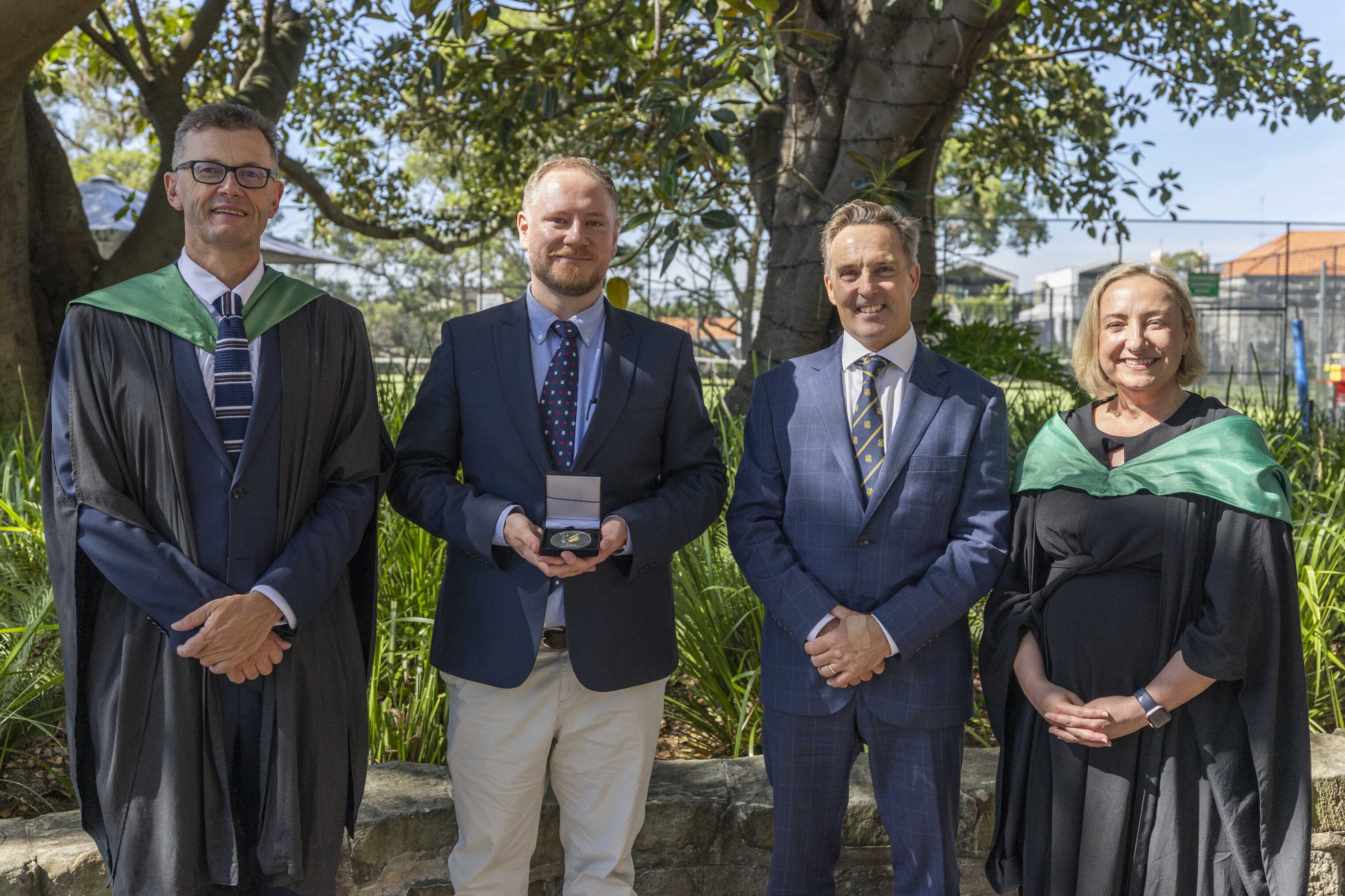 Four adults stand outdoors in formal attire, two in academic gowns with green sashes, one holding an award medal, all smiling in front of trees and a stone wall on a sunny day.