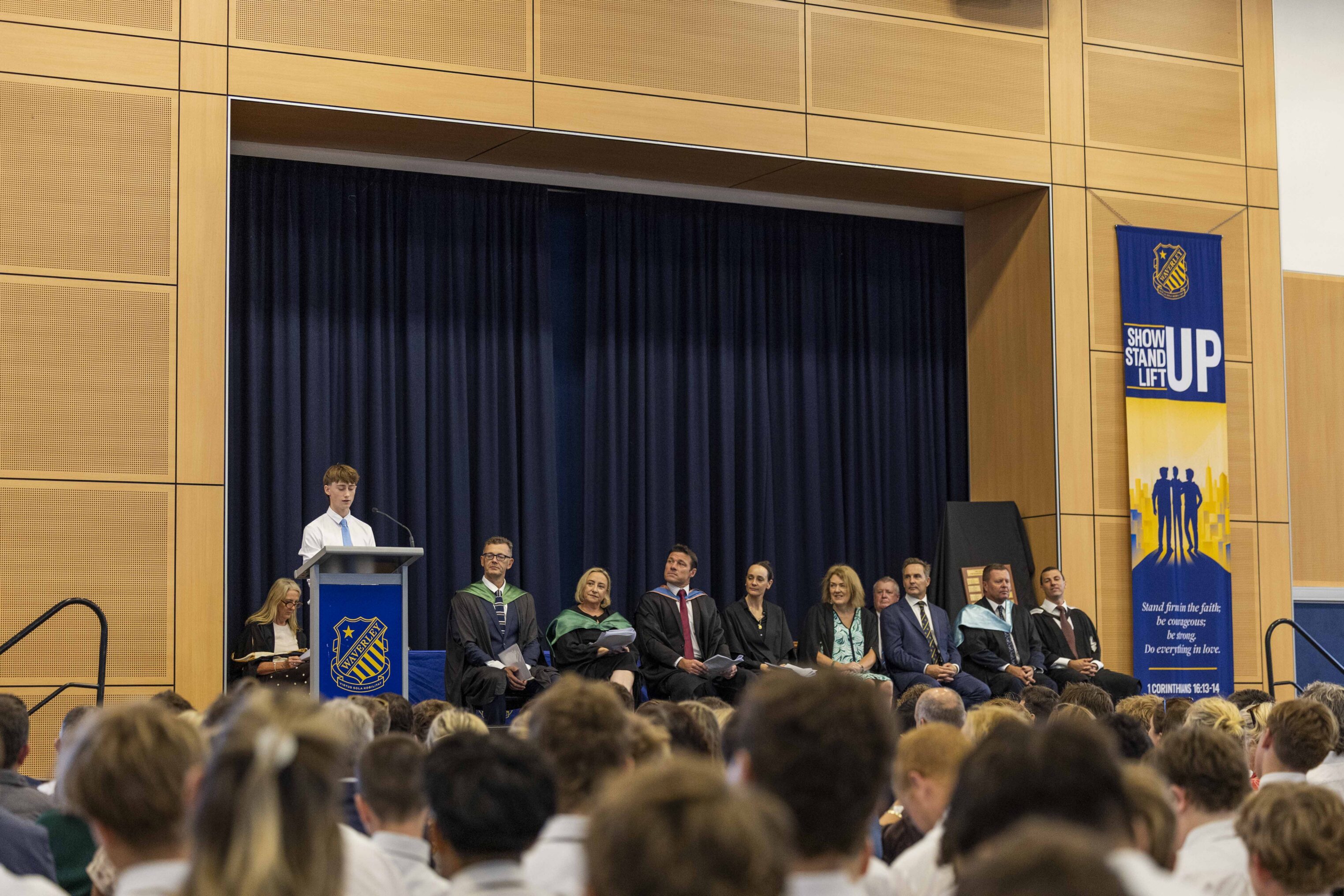 A student speaks at a lectern on stage in a school assembly hall. Several adults and pupils sit behind him, and a large banner reading “STAND UP” is displayed on the right. The audience consists of students in uniform.