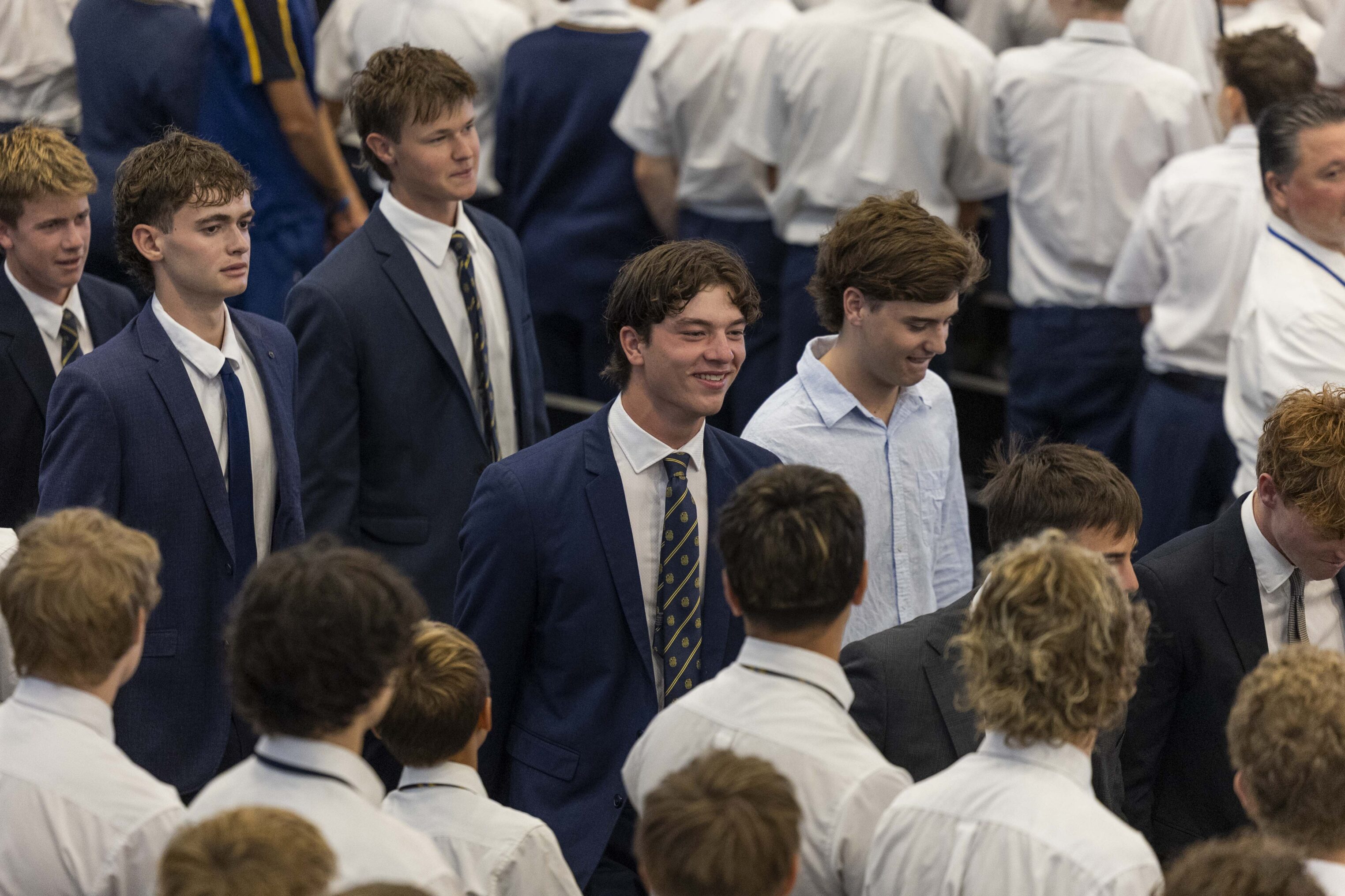 A group of young men, mostly dressed in white shirts and ties, stand closely together in a crowded indoor setting. One person near the centre wears a navy suit and striped tie, smiling whilst talking to others.
