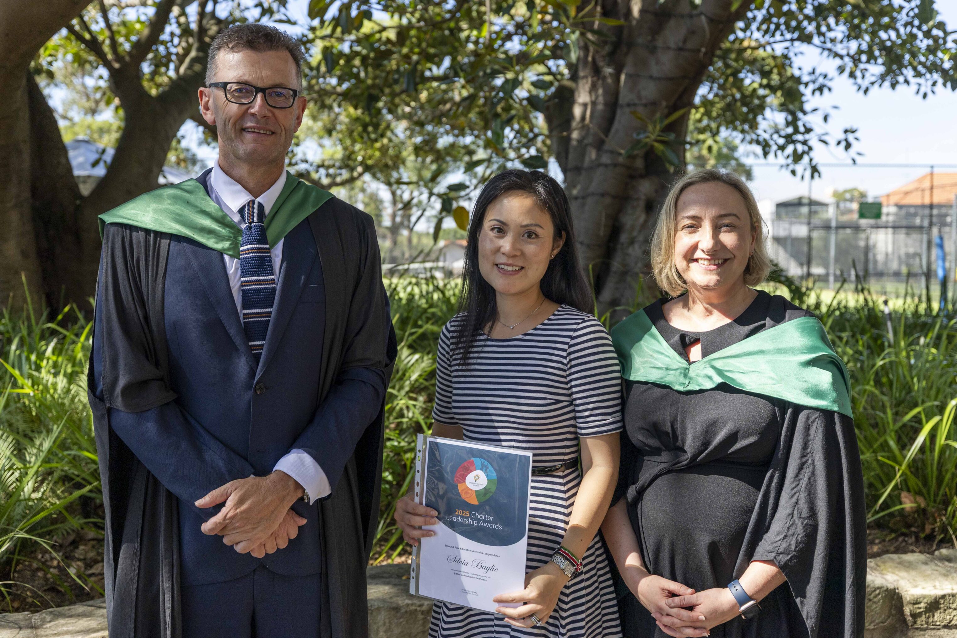 Three adults stand outdoors in graduation attire. The man on the left and woman on the right wear black gowns with green stoles. The woman in the centre, holding a certificate, wears a striped dress. Trees and greenery are in the background.