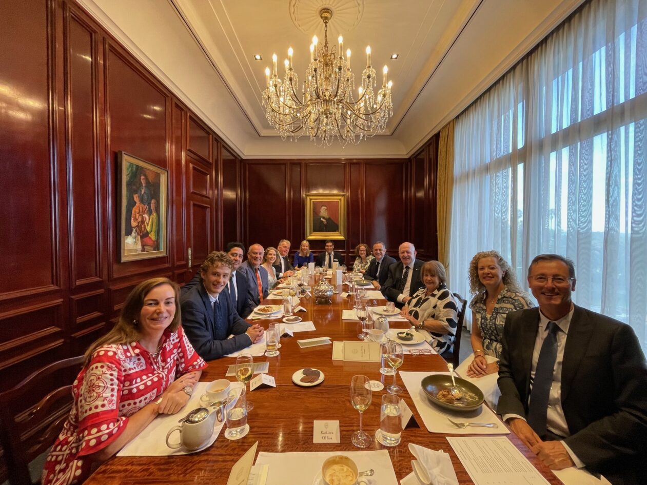 A group of people in formal attire sit around a long dining table in an elegant, wood-panelled room with a chandelier, large windows, and a framed portrait on the wall. Food and drinks are set on the table.