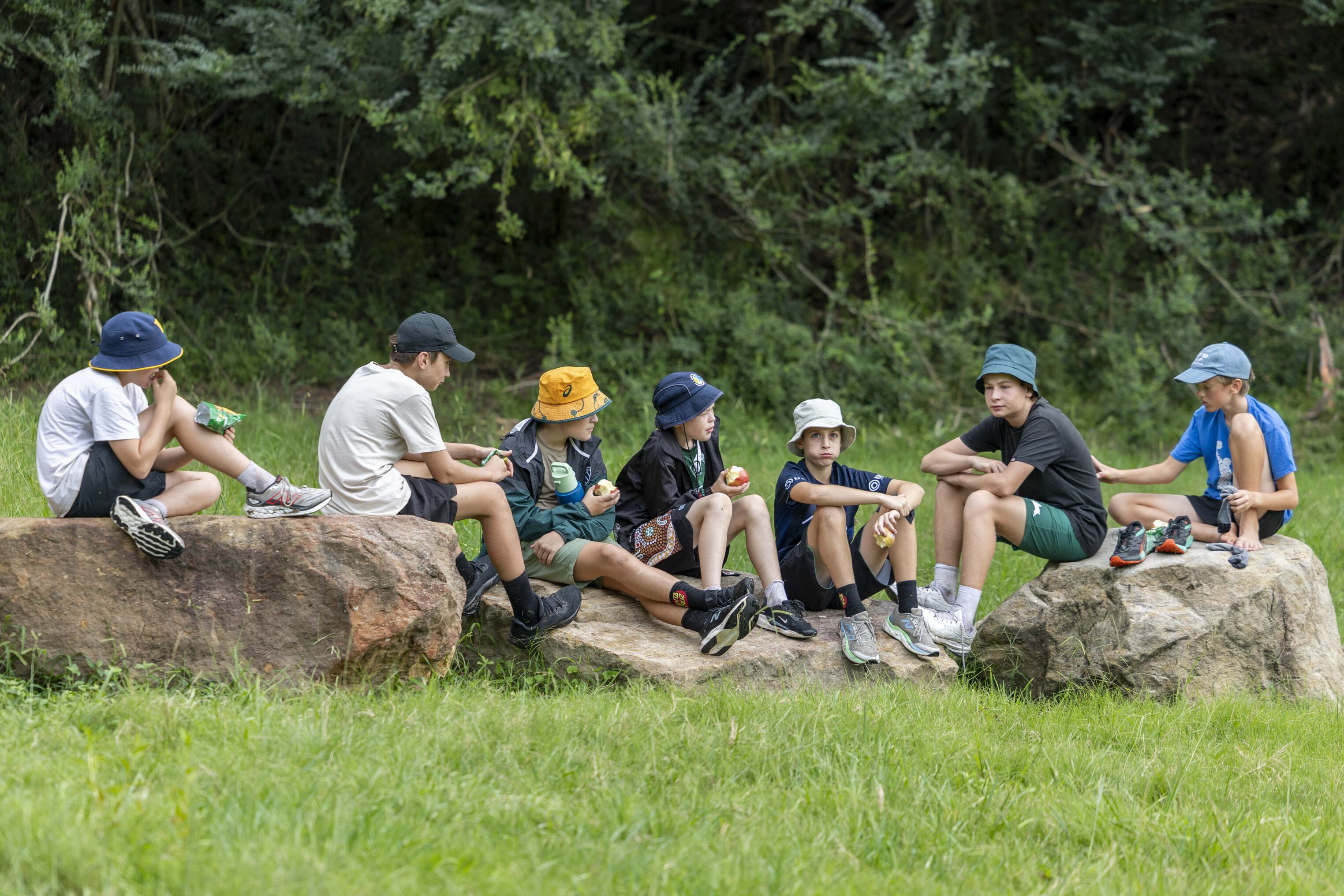 Seven children wearing hats sit on large rocks outdoors, talking and eating snacks. They are surrounded by green grass and trees, with an informal, relaxed atmosphere.