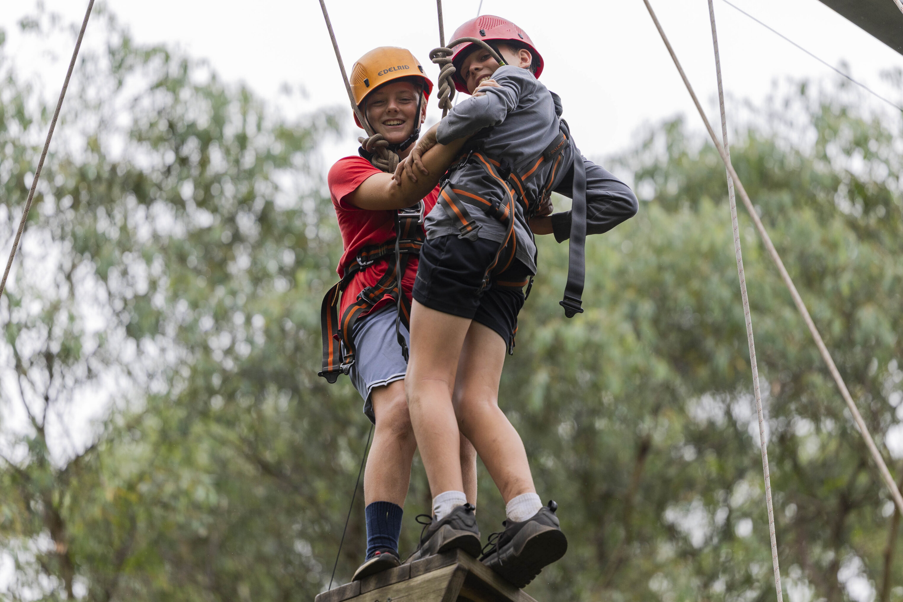 Two children wearing helmets and harnesses balance together on a wooden beam high above the ground, holding onto each other during an outdoor ropes course with trees in the background.