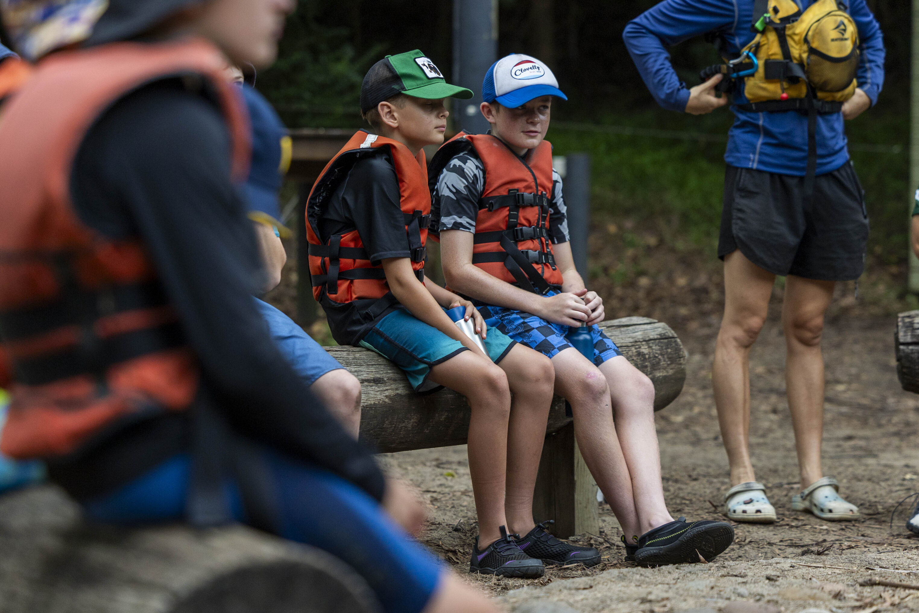 Two children wearing life jackets and baseball caps sit on a wooden log outdoors, listening attentively. An adult stands nearby in a blue jacket and shorts, with trees and soil in the background.