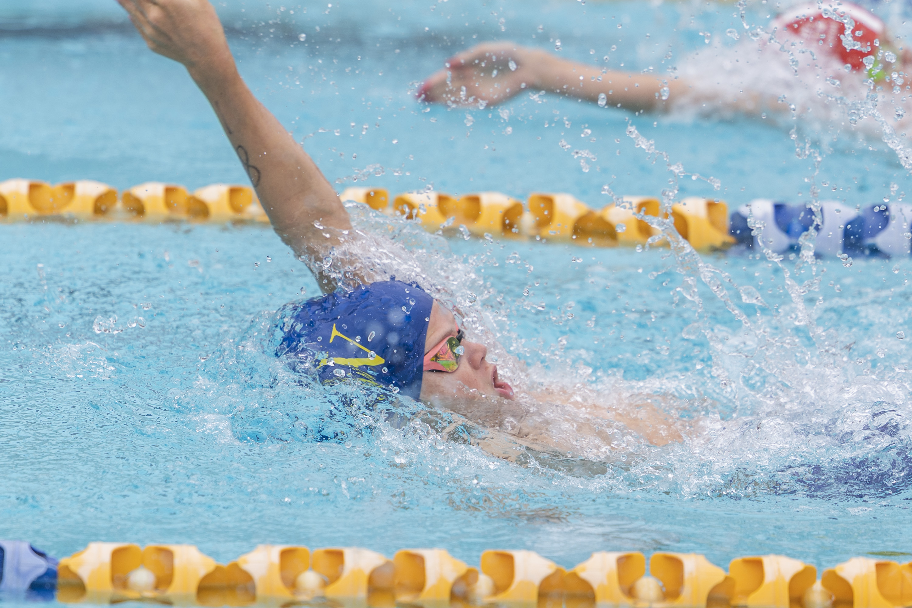 A swimmer wearing a blue cap and goggles performs the backstroke in a pool, surrounded by yellow lane dividers, with water splashing around them.