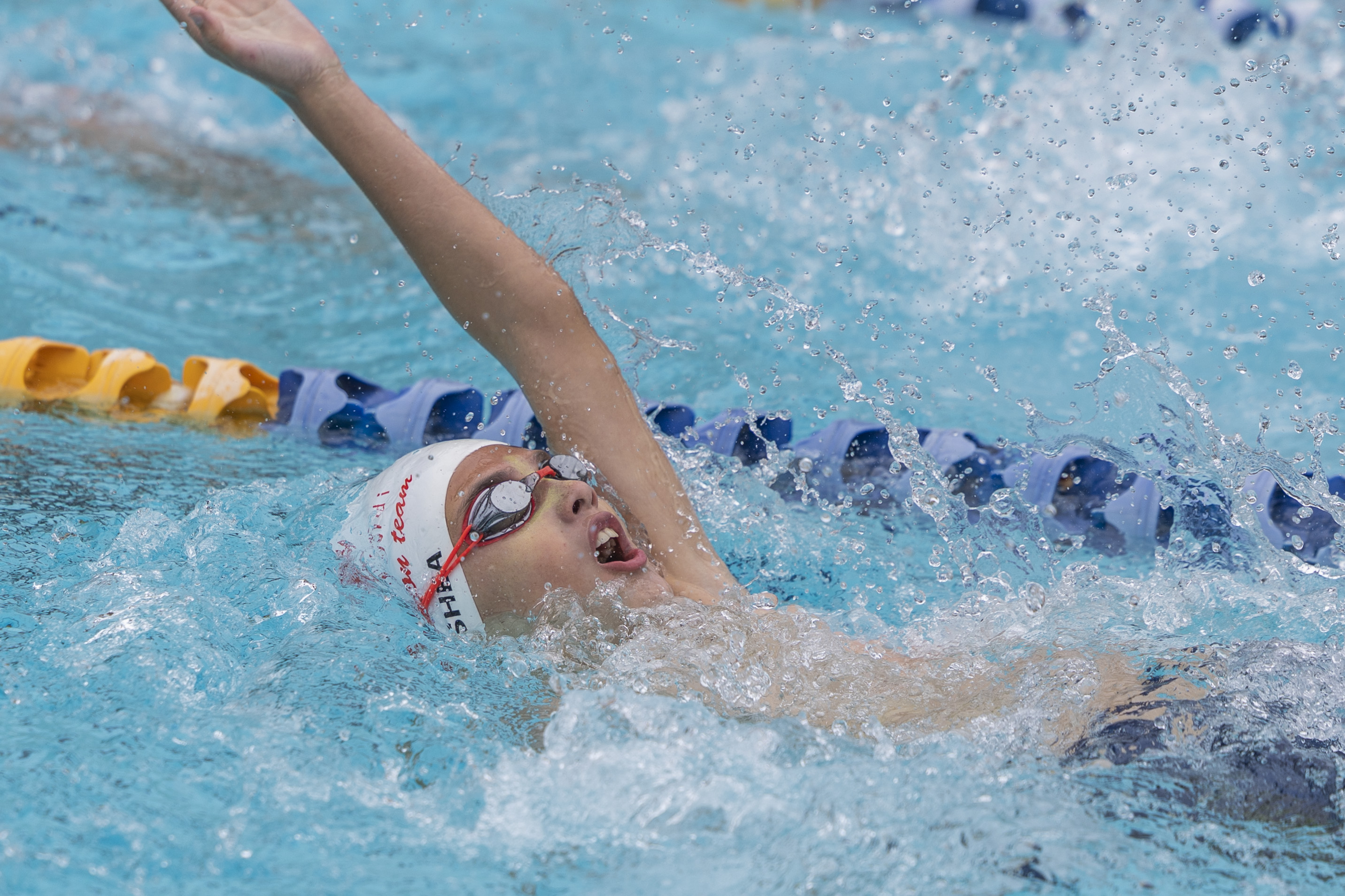 A young swimmer wearing a white swimming cap and goggles swims backstroke in a pool, raising one arm out of the water. Splashes surround the swimmer, and lane dividers are visible in the background.