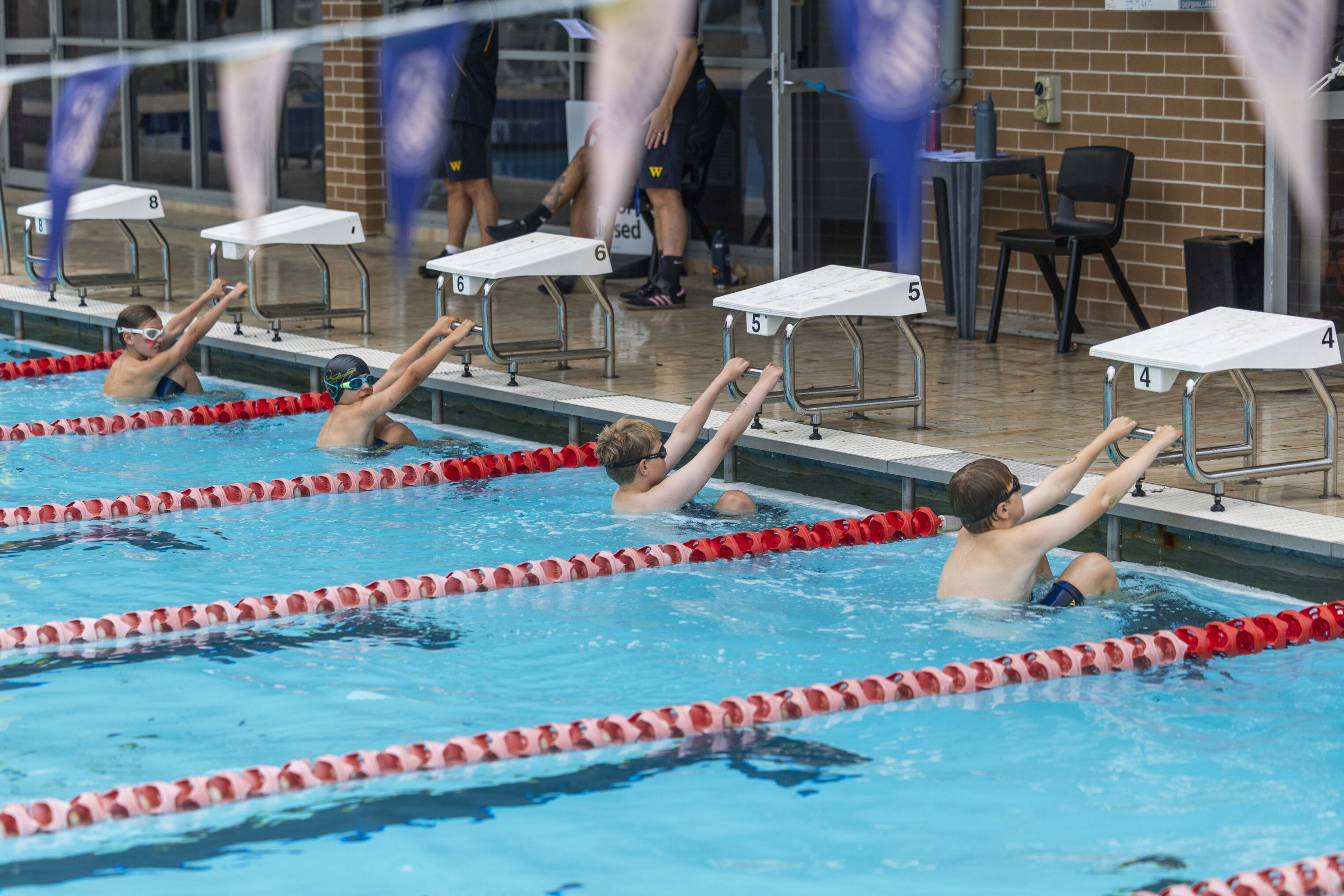 Four young swimmers in goggles and swim caps prepare for a backstroke race at an indoor pool, holding onto the wall at their respective lanes, with officials and empty chairs in the background.