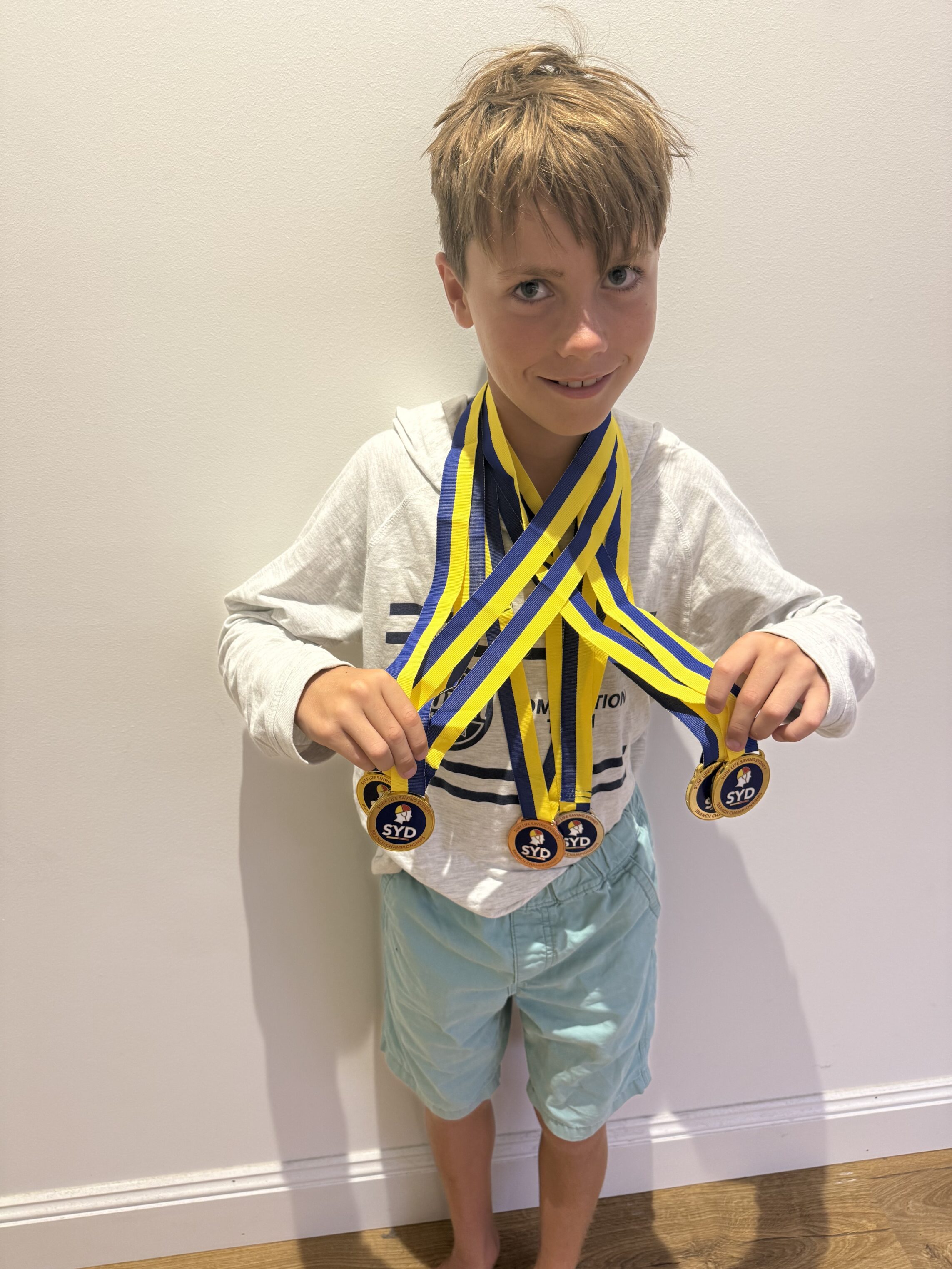 A young boy with light brown hair stands against a plain wall, smiling and wearing a white hoodie and light blue shorts. He holds four medals with blue and yellow ribbons round his neck.