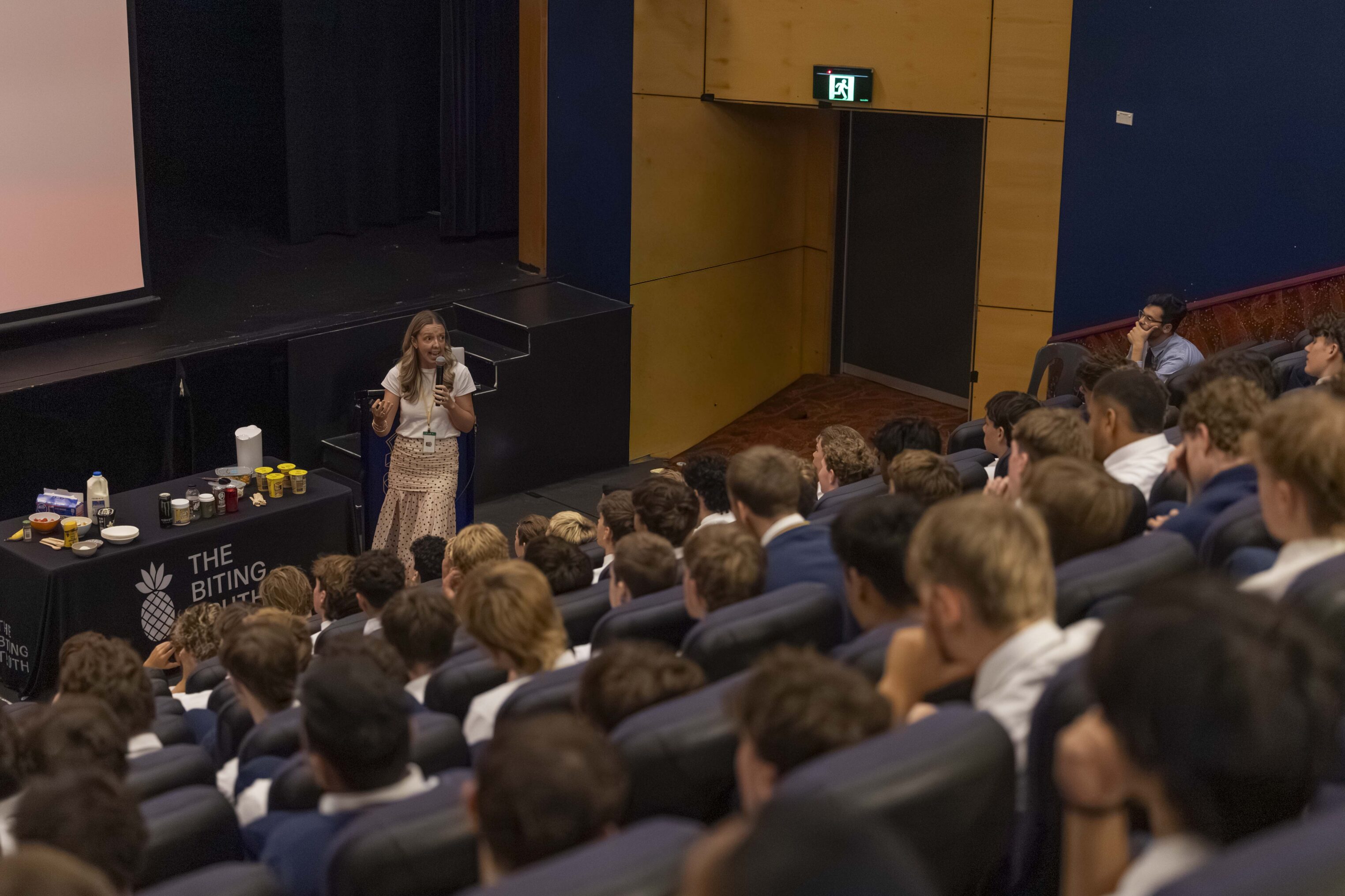 A woman stands on stage speaking to an audience of students seated in an auditorium. A table beside her holds various items, including containers and bottles. The students are wearing uniforms.