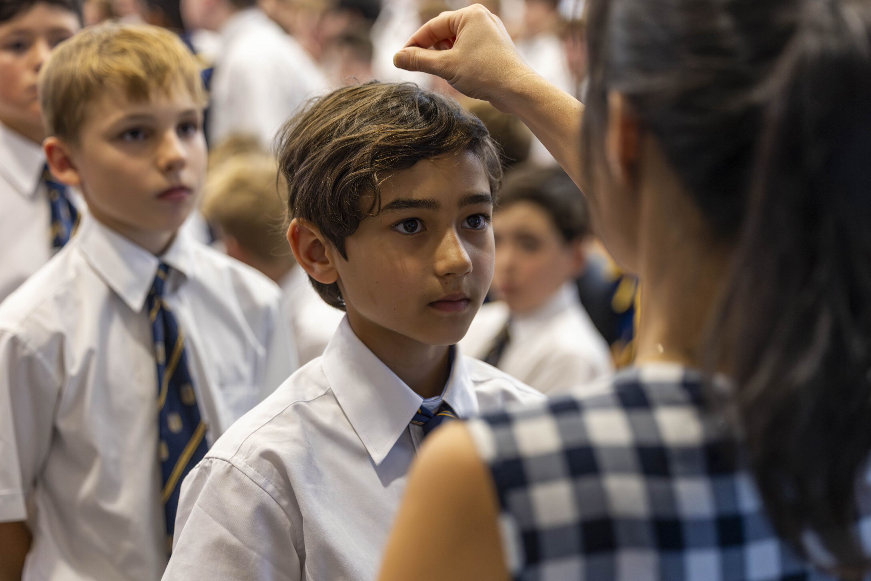 A woman faces a boy in a white shirt and tie, raising her hand as if marking his forehead. Other children in similar uniforms stand nearby, looking on. The setting appears to be a school event or ceremony.