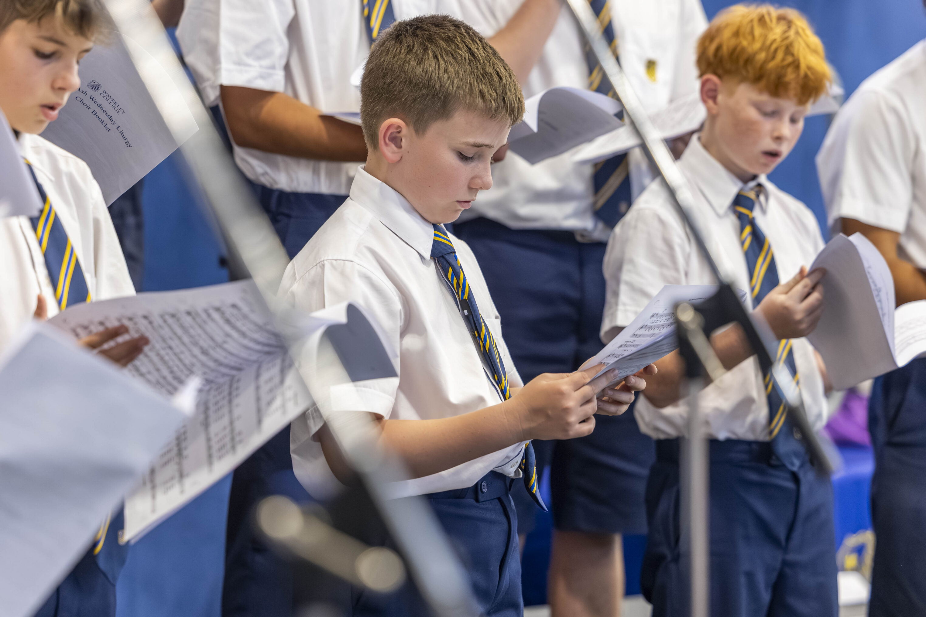 A group of boys in school uniforms and ties sing from sheets of music, standing close together in a choir setting, with microphones visible in the foreground.