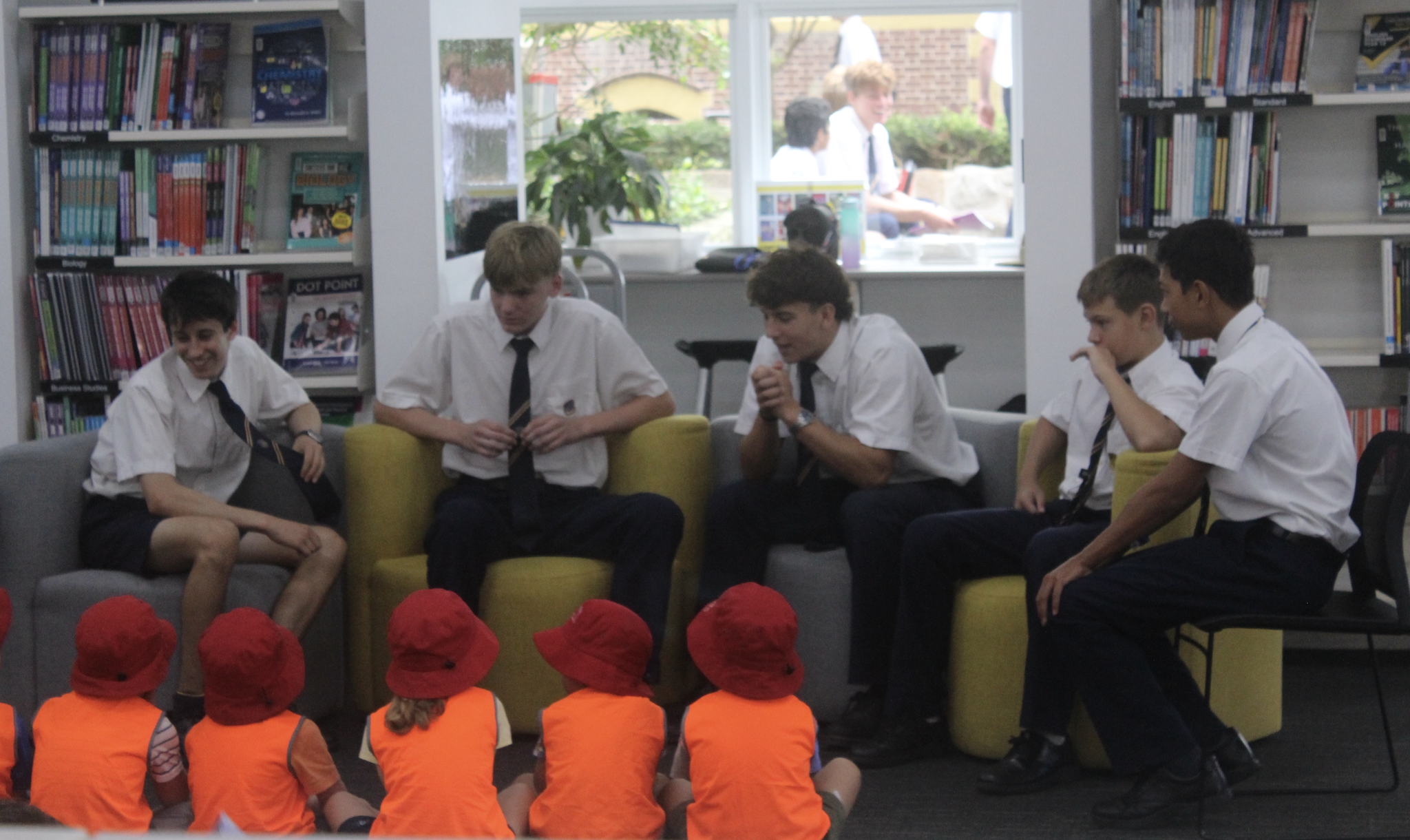 Five older students in school uniforms sit on chairs, speaking to a group of young children wearing orange shirts and red hats, in a library setting with bookshelves and a window in the background.