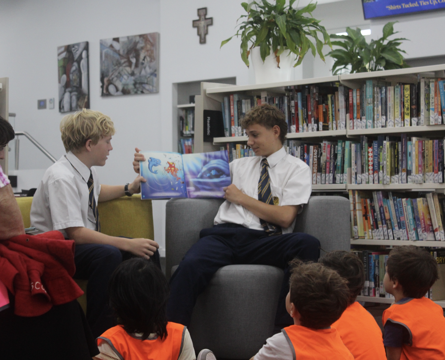 Two boys in school uniforms sit in a library, one holding up a colourful picture book and reading to a group of young children in orange vests seated on the floor, surrounded by bookshelves and plants.