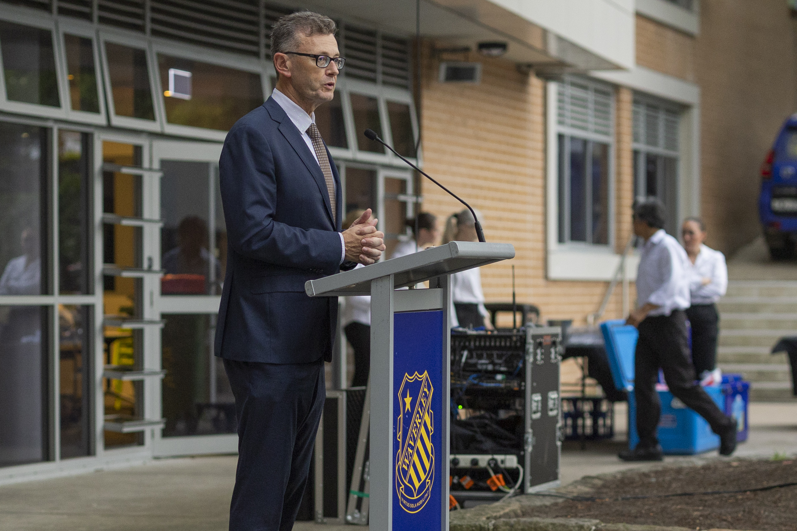 A man in a suit and glasses speaks at a lectern outdoors near a building, with people and equipment visible in the background. The lectern has a blue shield logo on the front.