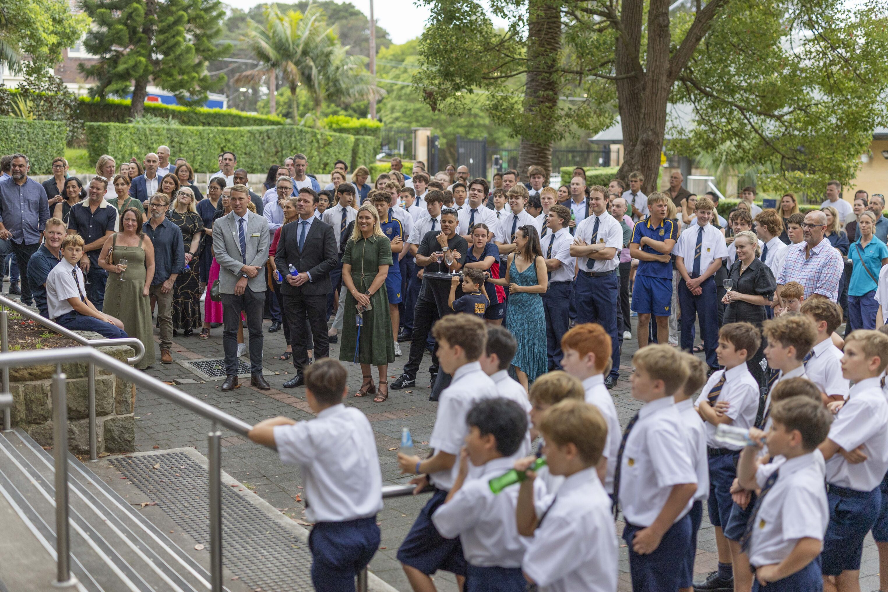 A large group of adults and children, many in school uniforms, gather outdoors near steps, watching something ahead. Trees and greenery surround the area, giving a lively, community event atmosphere.