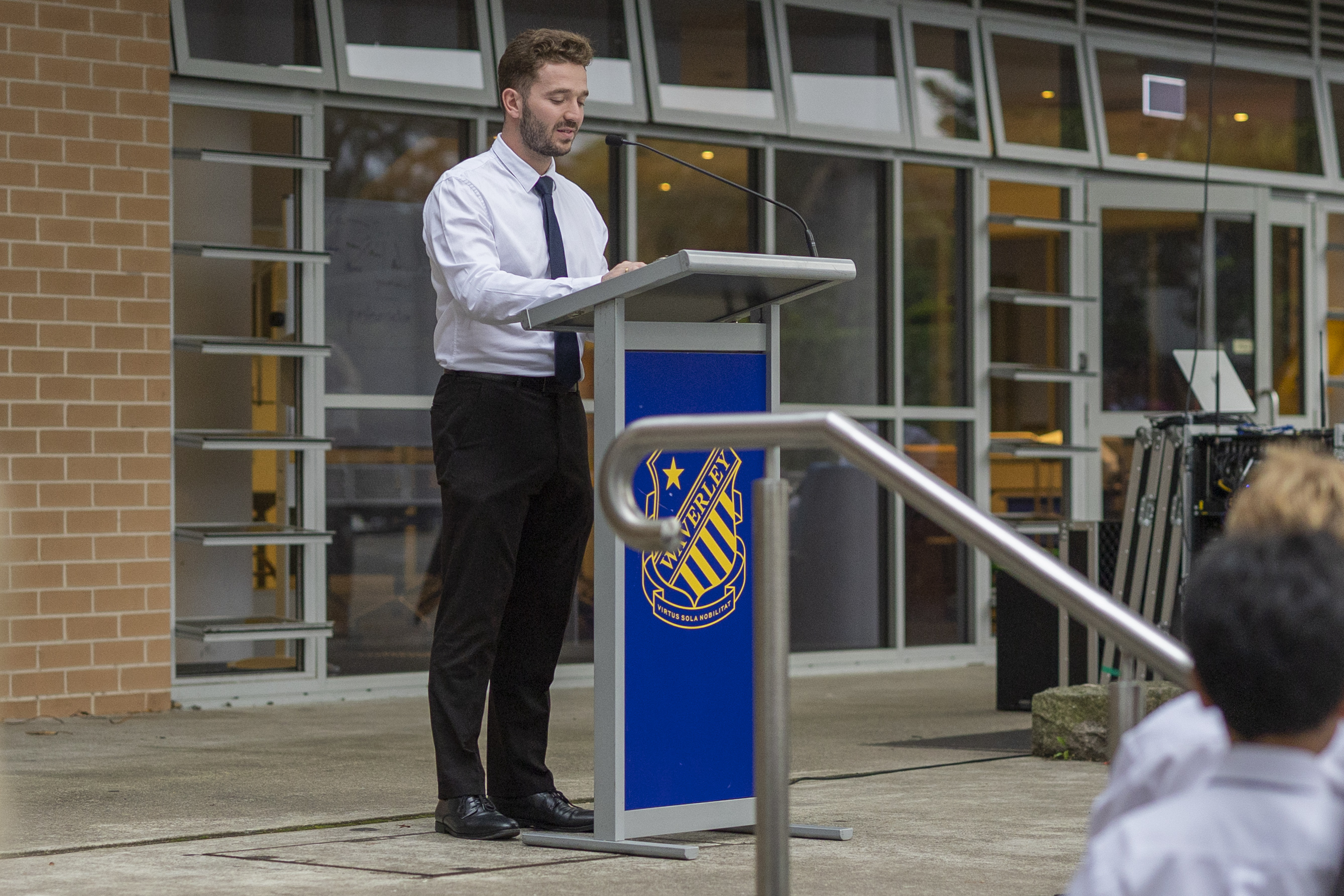 A man in a white shirt and black tie speaks at a lectern with a blue emblem outside a building with large windows, whilst people seated in front listen.