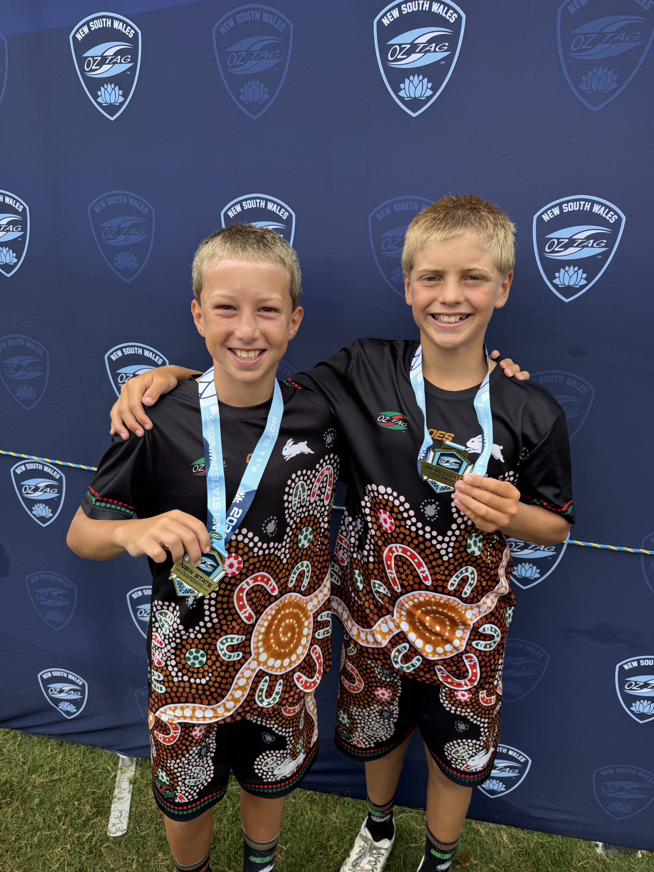 Two smiling boys in matching shirts with colourful Aboriginal designs stand arm in arm, each holding a medal. They pose in front of a blue New South Wales Oztag event backdrop.