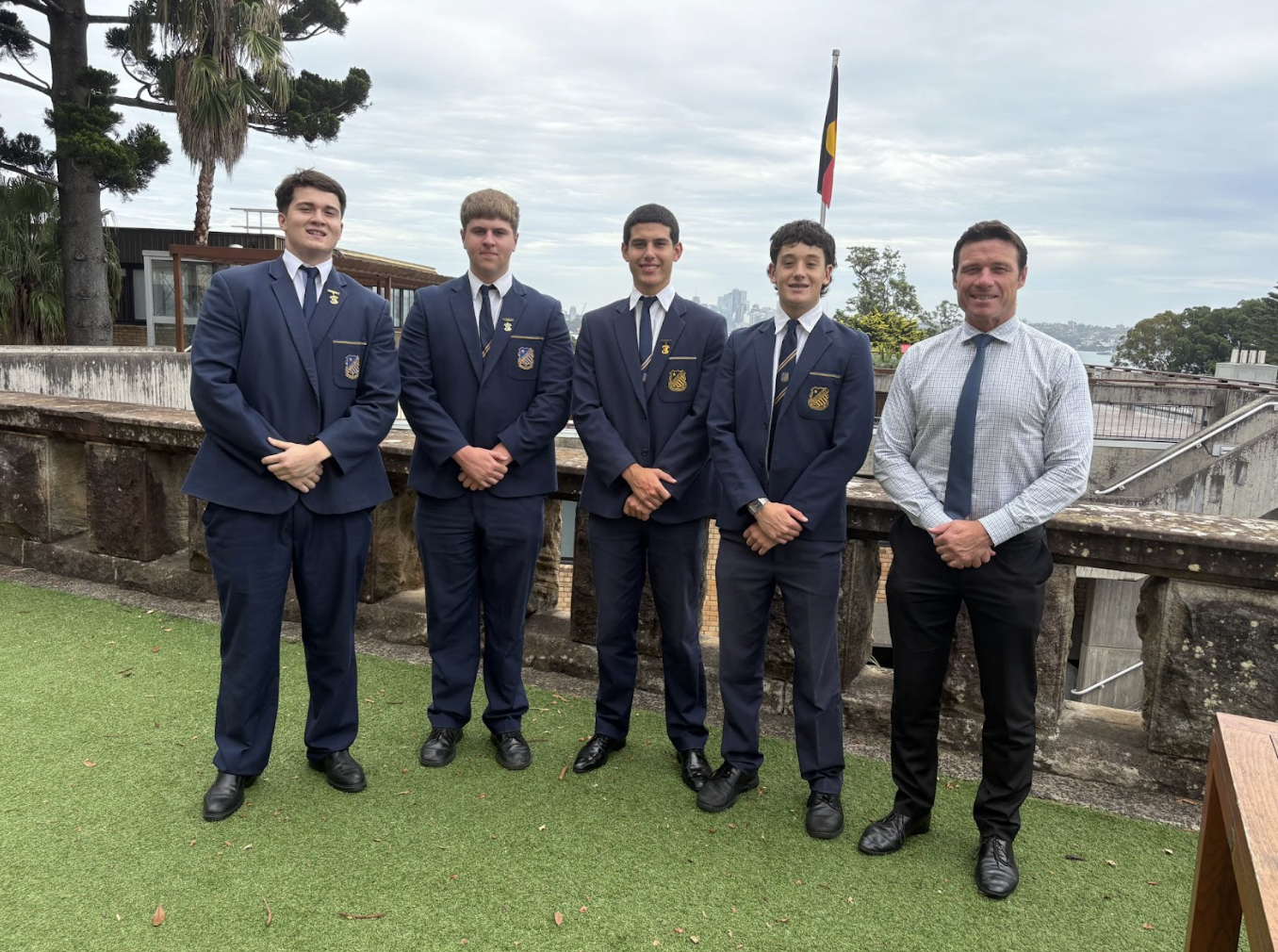 Five men stand in a row outdoors on grass, four wearing matching navy blue school uniforms and one in a white shirt and tie. A stone wall, trees, and a flag are visible in the background.