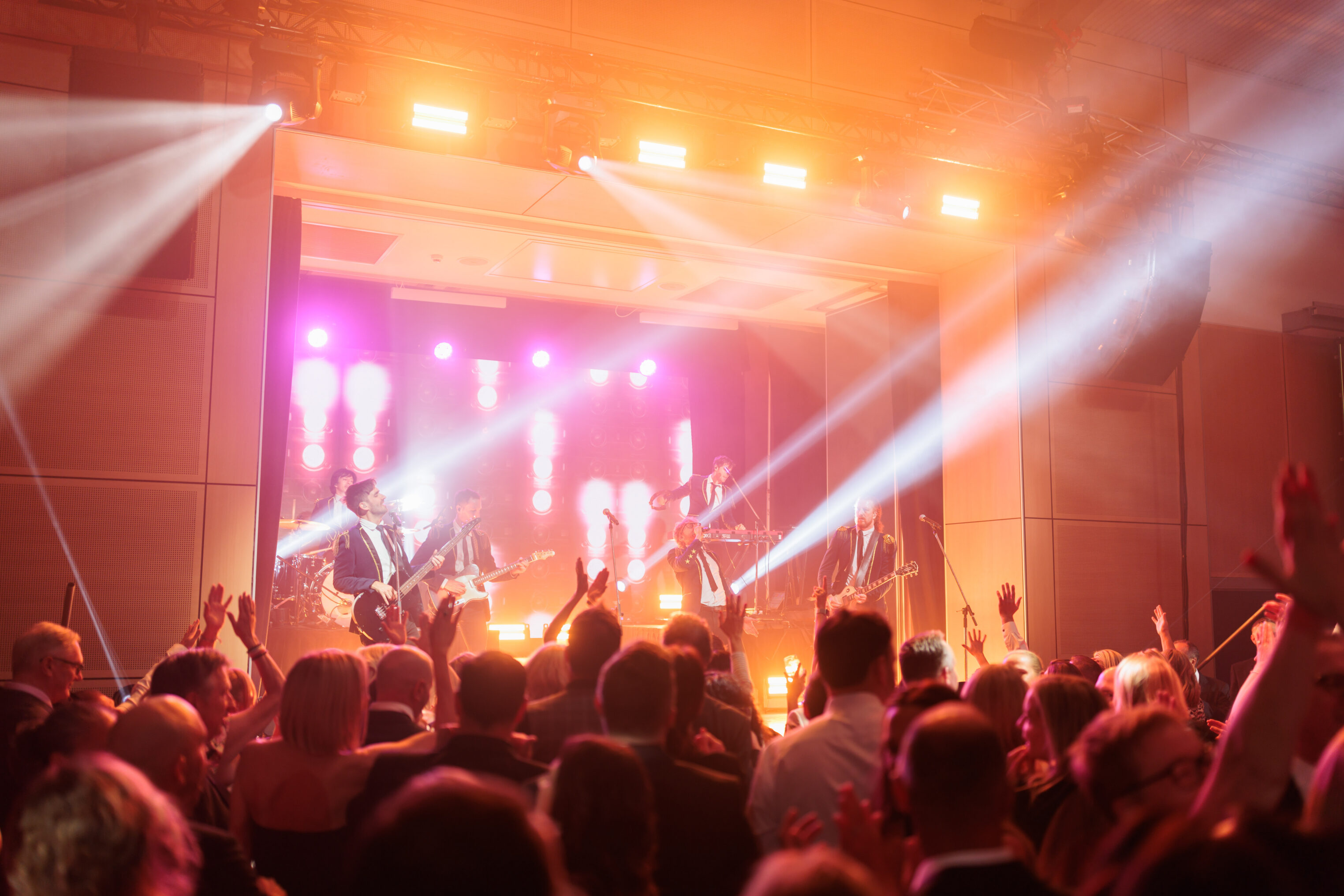 A lively band performs on stage under bright lights, with a large crowd of people enjoying the concert in an indoor venue. Audience members raise their hands and the atmosphere is energetic and festive.