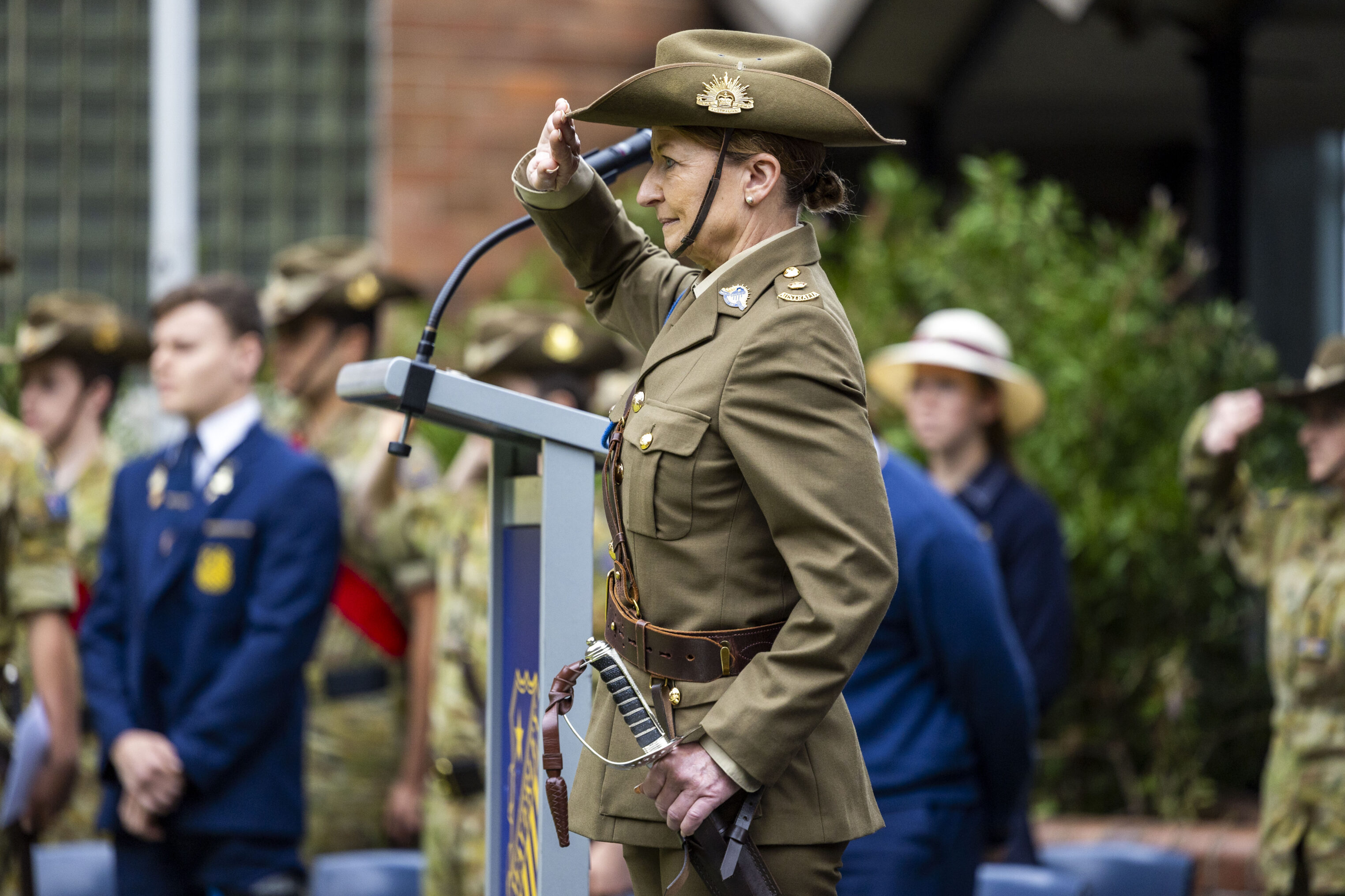 A uniformed soldier stands at a lectern, saluting, during a ceremony. Other soldiers and attendees are visible in the blurred background.