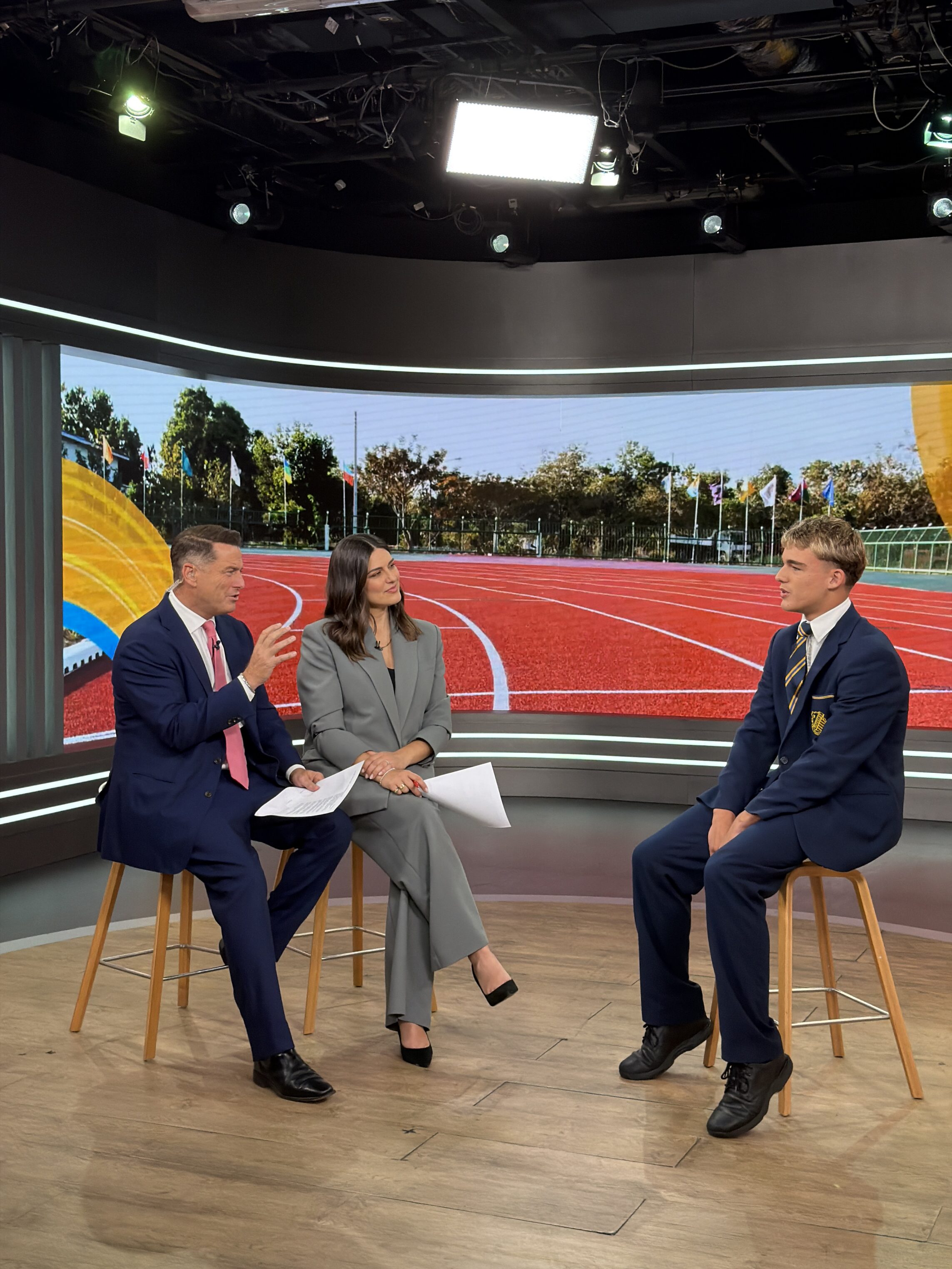 Two TV presenters in suits interview a young man in a school blazer on a set with a large screen displaying an athletics track and stadium background. All three are seated on stools, engaged in conversation under studio lights.