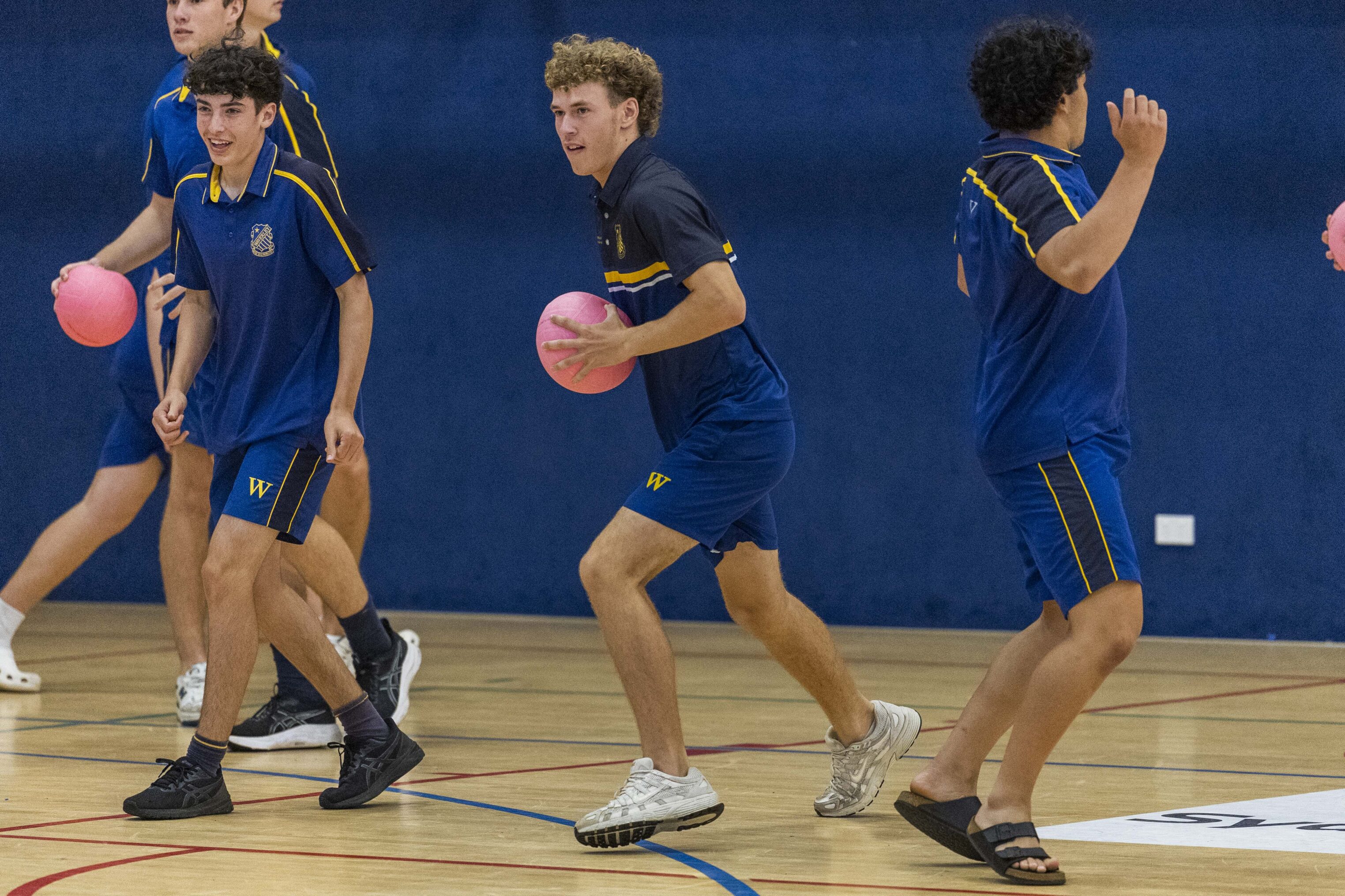 Four young men wearing blue sports kits play dodgeball indoors. Two are holding pink balls, and all appear to be moving or preparing to throw. The gym has a wooden floor and dark blue walls.