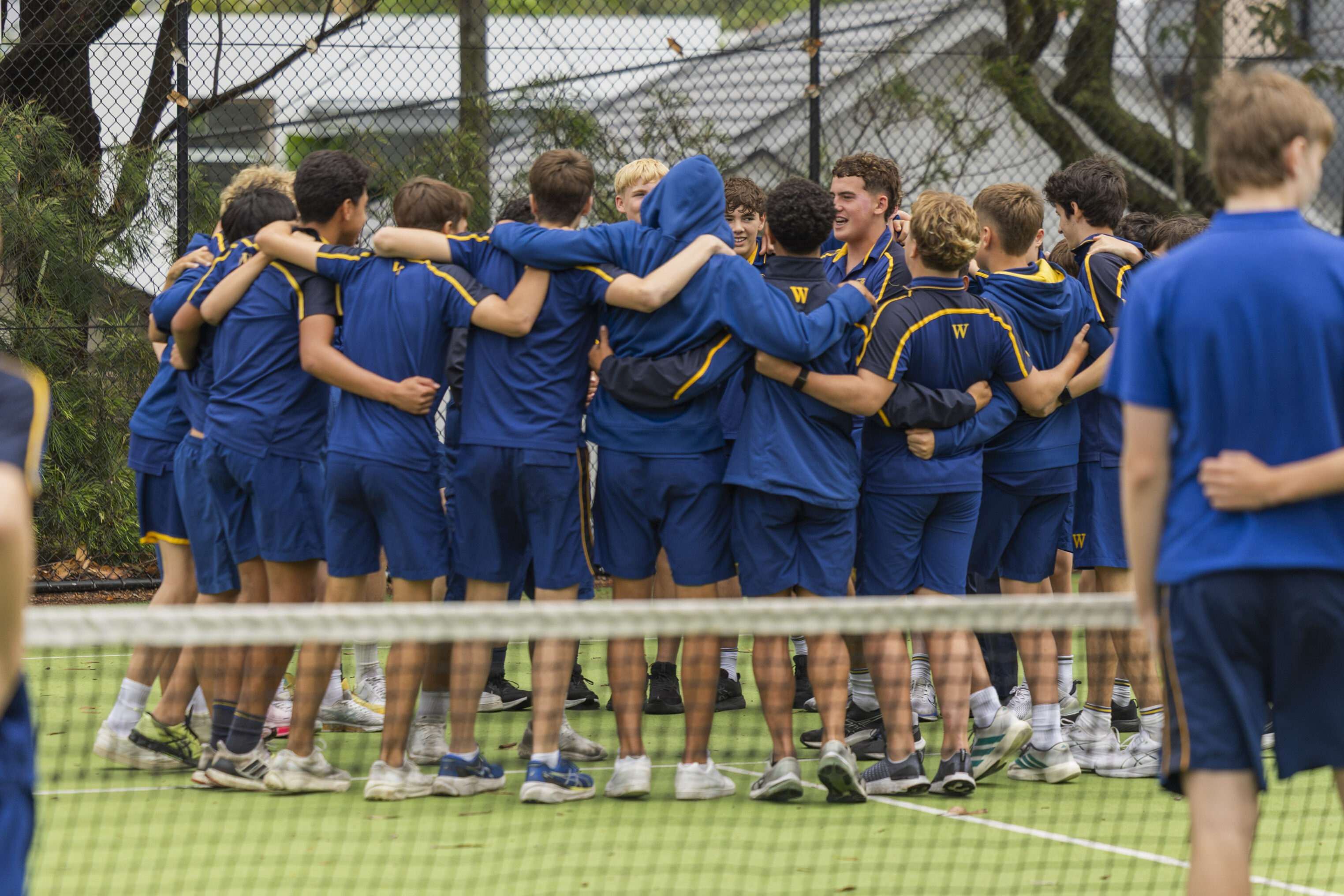 A group of teenage boys in blue sports kits stand in a circle with arms round each other on a tennis court, appearing to cheer or motivate each other before a match.