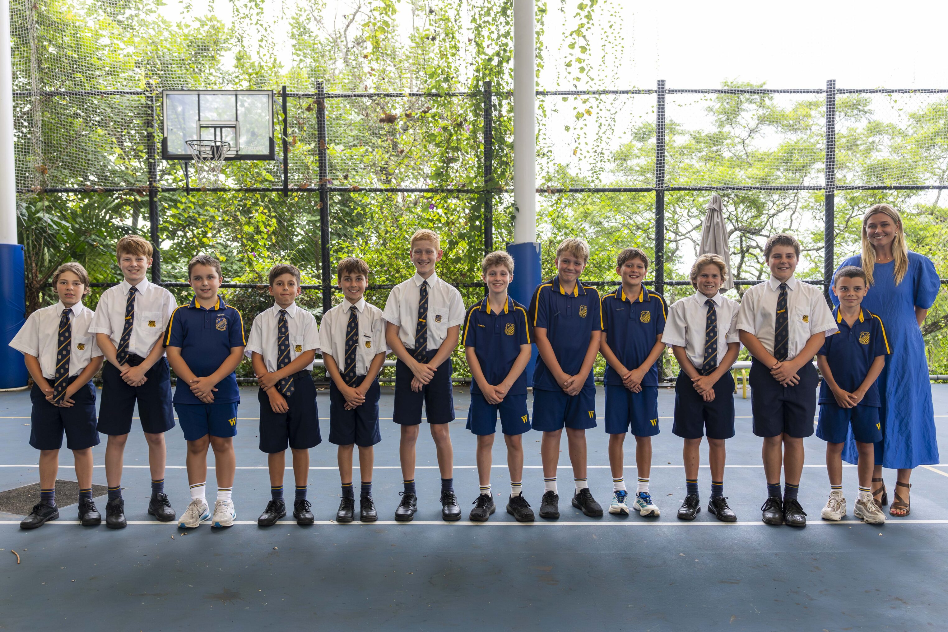 A group of twelve boys and one woman stand in a line on a basketball court. The boys wear school uniforms in white shirts or blue sports kit, and the woman wears a blue dress. Trees and greenery are visible in the background.