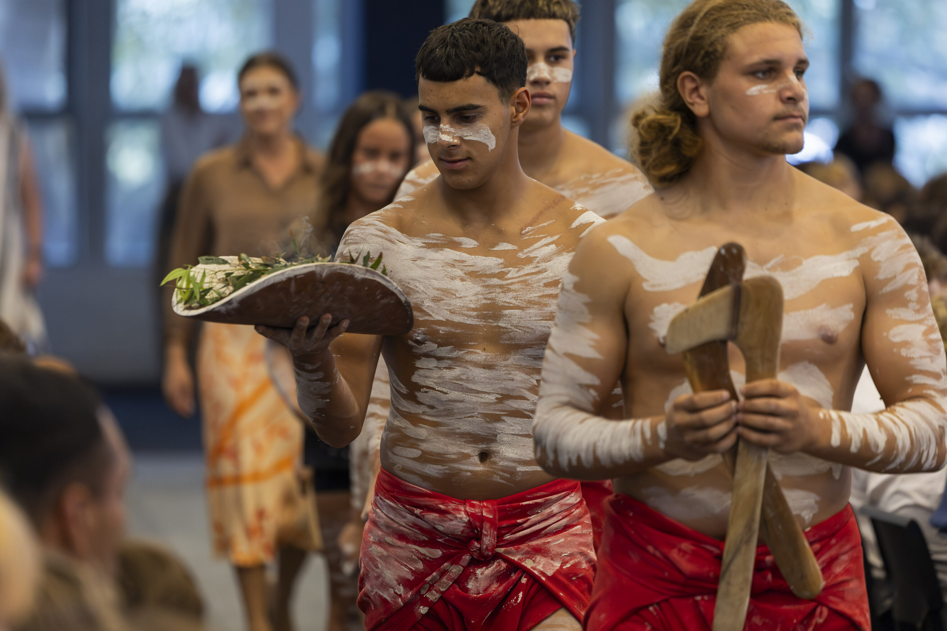 Three young men with traditional white body paint and red cloths participate in a cultural ceremony indoors. One carries a wooden platter with leaves; another holds wooden objects. Onlookers are seated in the background.