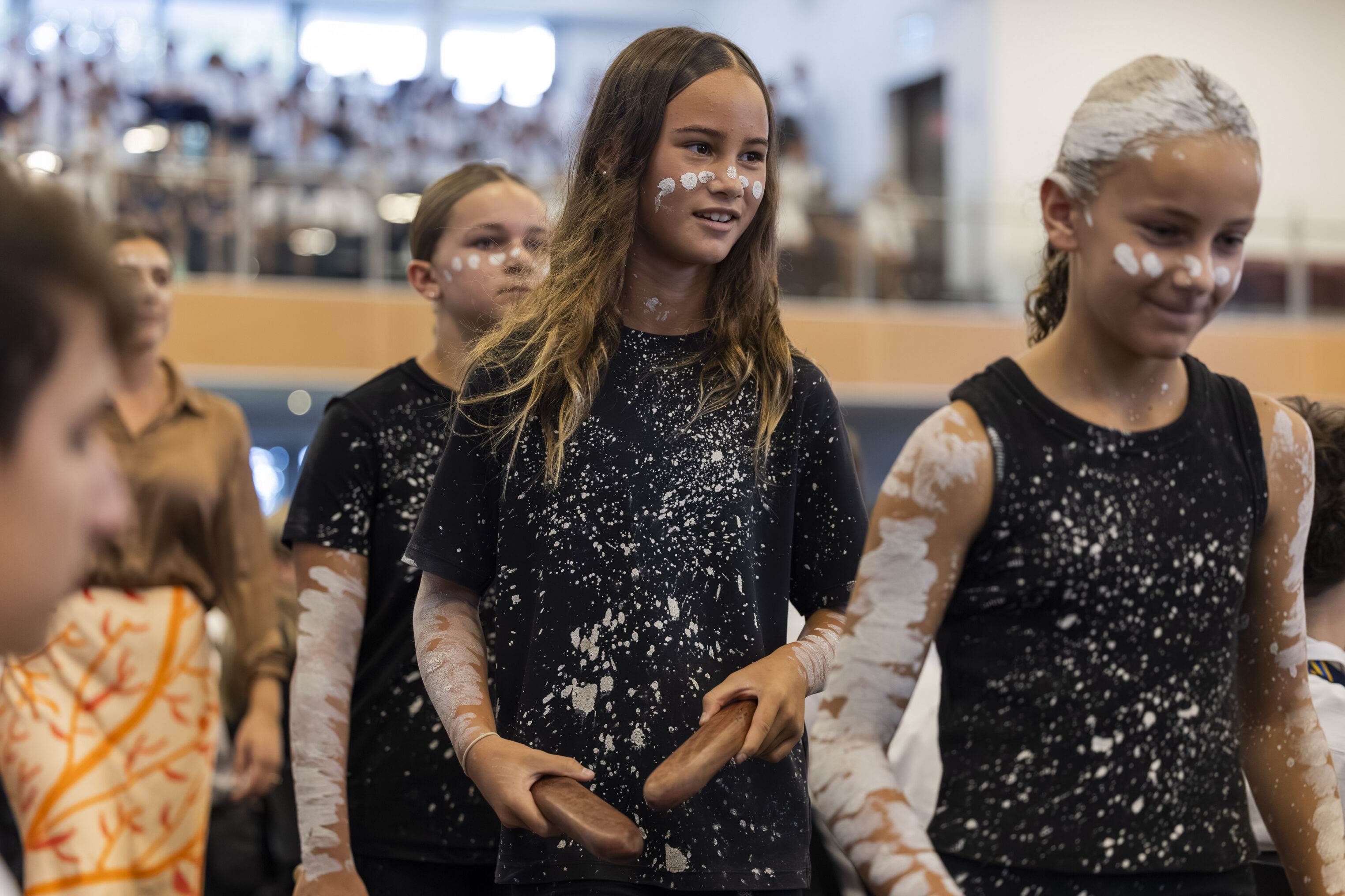 Three girls wearing black shirts with white paint splatters and traditional white face and arm paint participate in a cultural event indoors, with one holding two wooden clapsticks. Other people are visible in the background.