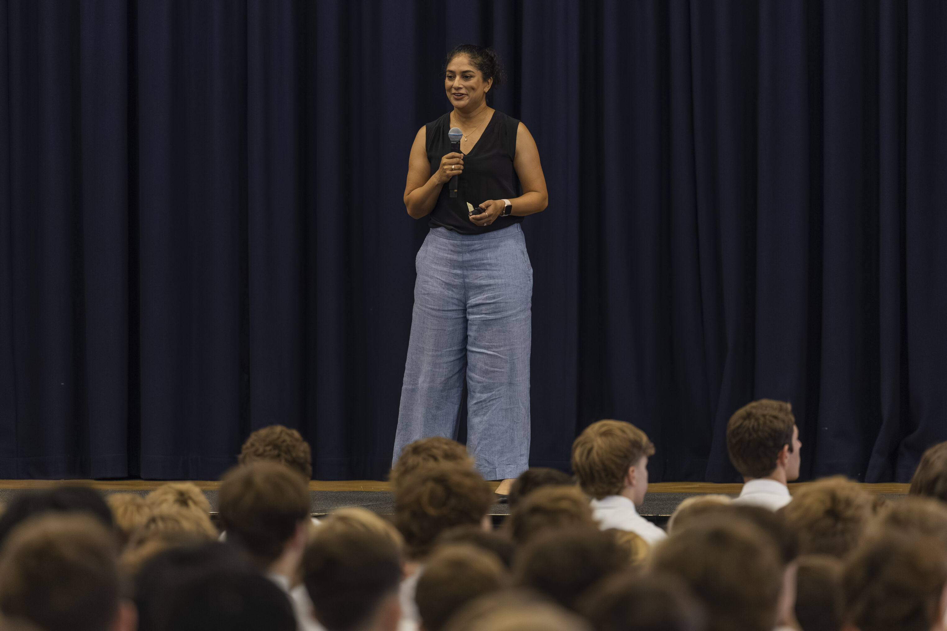 A woman holding a microphone stands on stage in front of a blue curtain, speaking to an audience of seated students in white shirts.