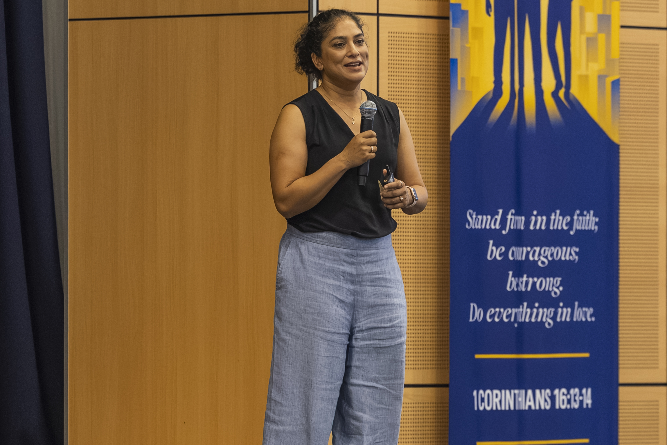 A woman stands holding a microphone and smiling, wearing a black sleeveless top and light blue trousers. Beside her is a banner with a Bible verse: “Stand firm in the faith; be courageous; be strong. Do everything in love.”.