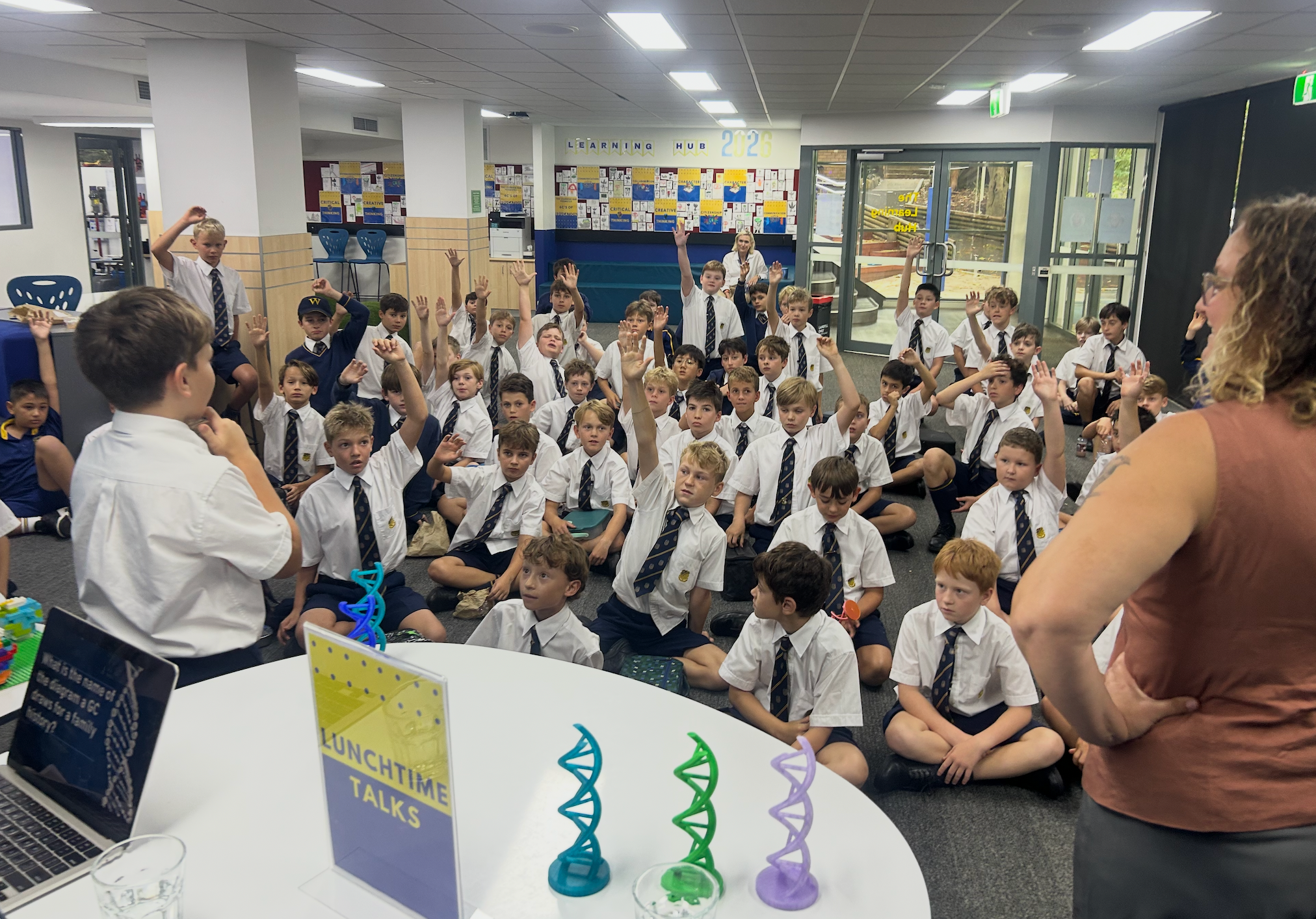 A large group of boys in school uniforms sit on the floor, some raising their hands, whilst a woman stands nearby. In the foreground is a table with colourful DNA models and a sign that reads “Lunchtime Talks”.