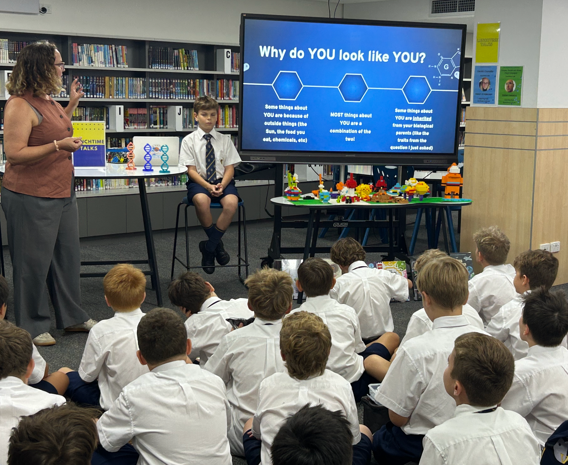 A woman stands beside a seated boy and a TV screen displaying Why do YOU look like YOU? whilst a group of schoolchildren in white shirts sit on the floor, watching in a library setting.