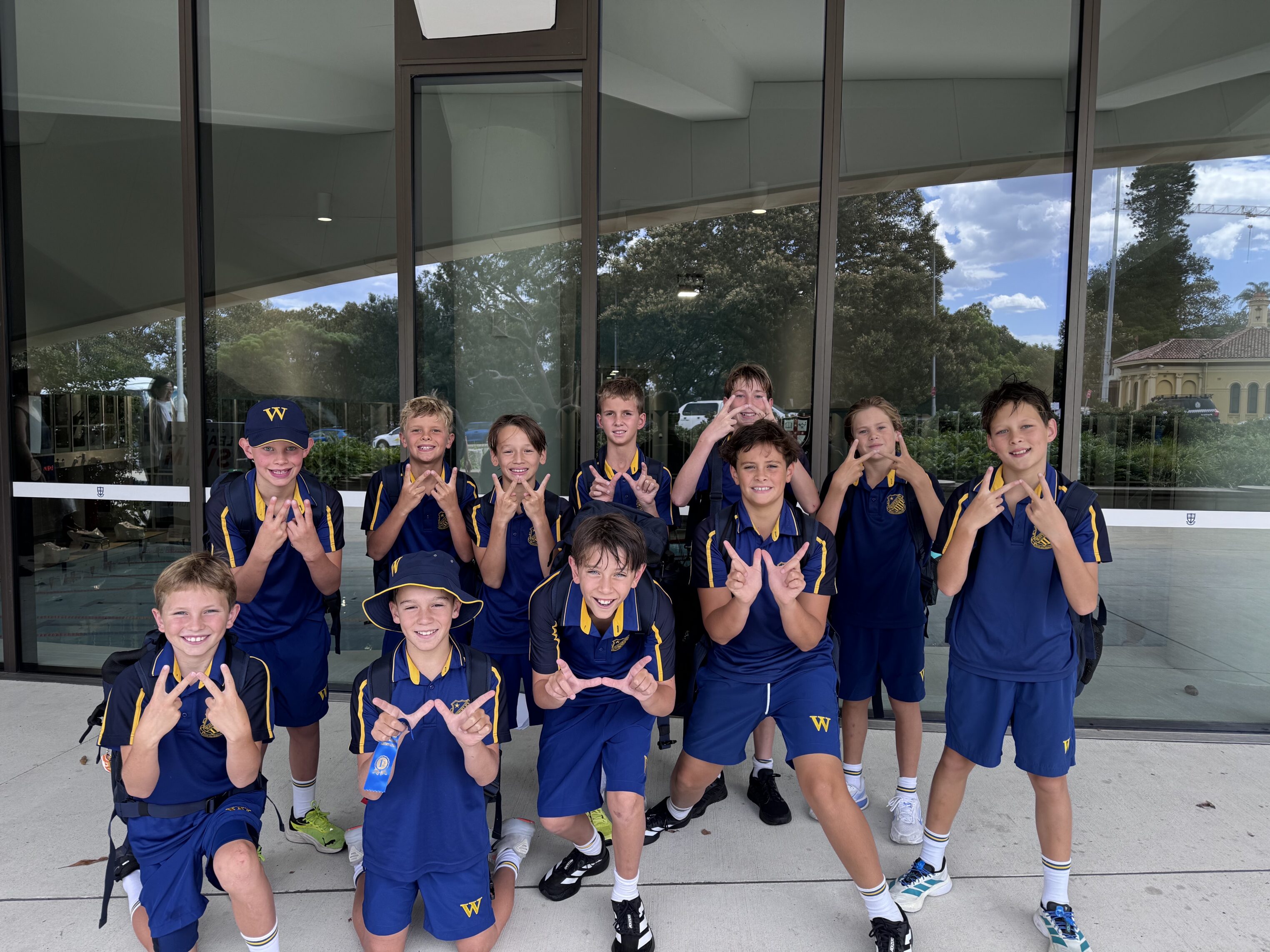 A group of ten boys in matching blue and yellow school uniforms pose outside a building, smiling and making W shapes with their hands. Trees and sky are reflected in the glass windows behind them.