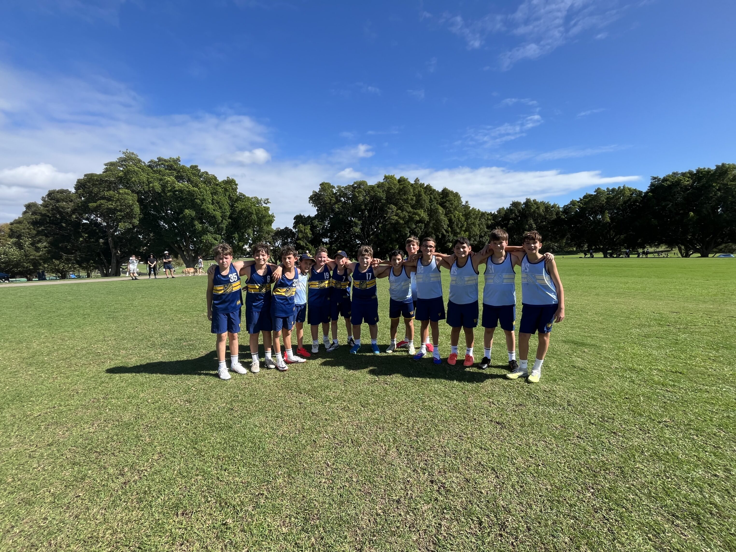 A group of teenage boys wearing blue and yellow sports kits stand together in two rows on a grassy field, smiling under a bright blue sky with scattered clouds and trees in the background.