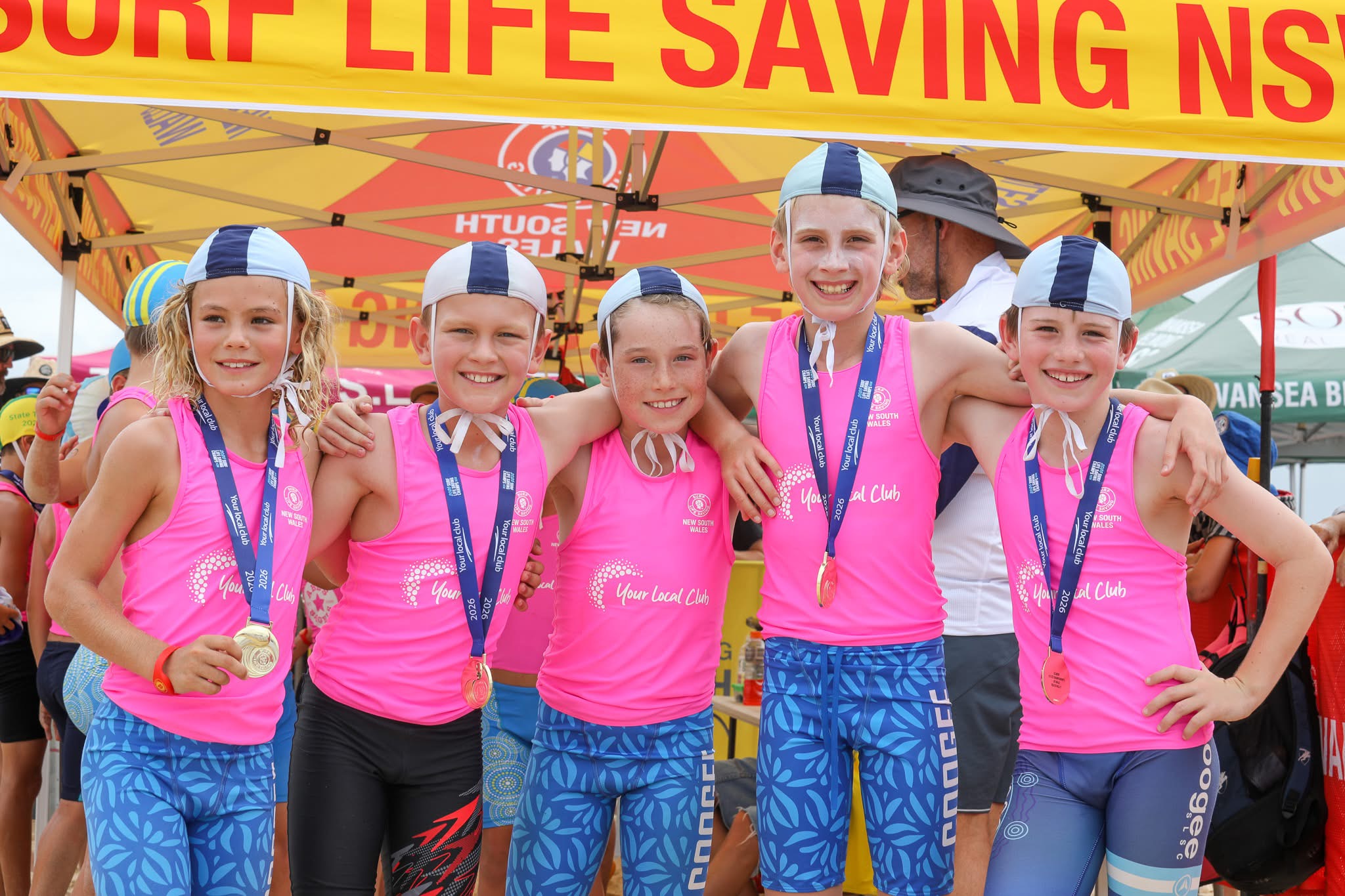 Five smiling children in matching pink shirts, blue swimming caps, and patterned blue shorts stand side by side under a Surf Life Saving tent, each wearing a medal. They have their arms round each other, celebrating together.