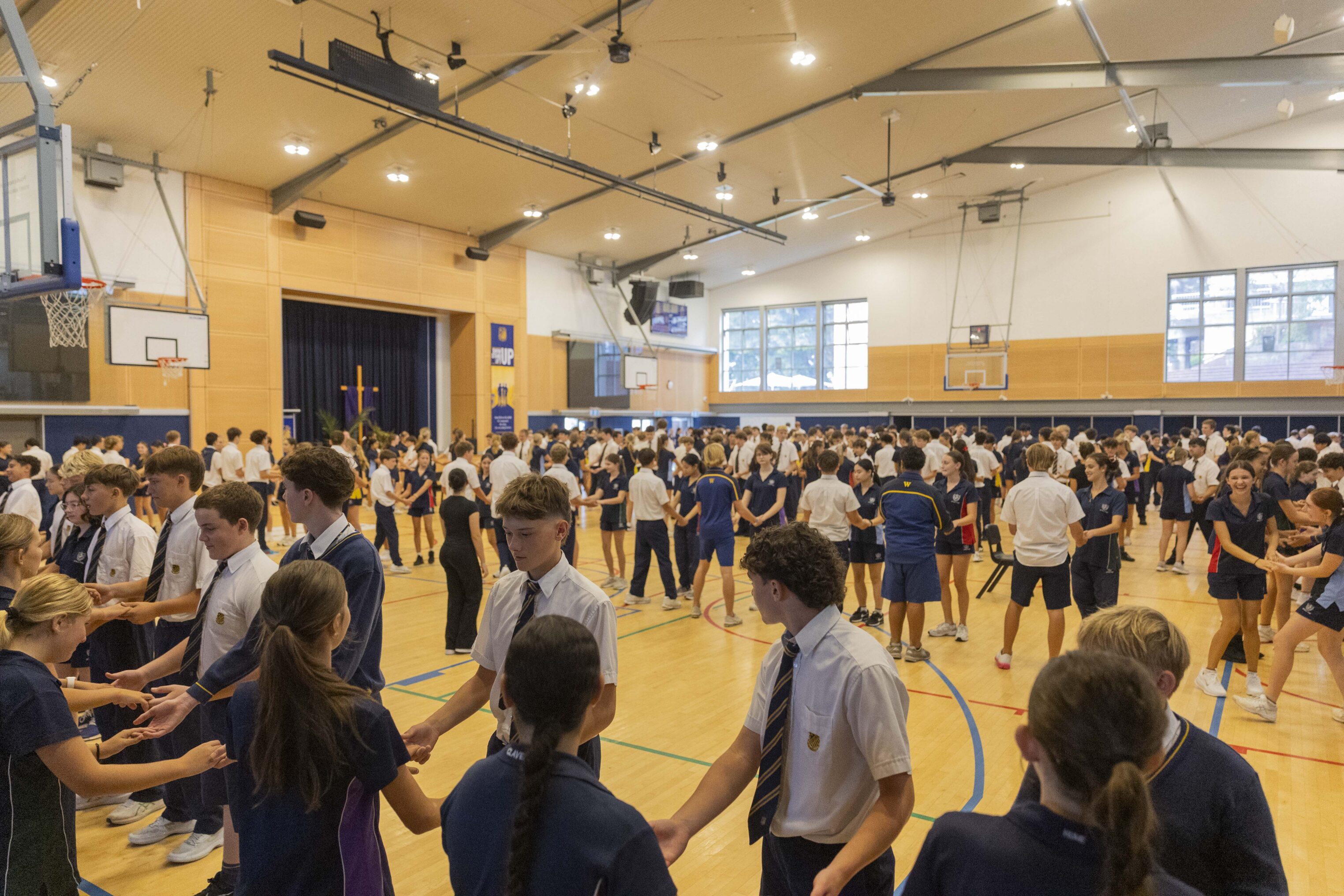 A large group of students gathered in a gymnasium, standing in pairs and holding hands. The students are dressed in uniforms and sportswear. The gymnasium is brightly lit with basketball hoops and wooden floors.