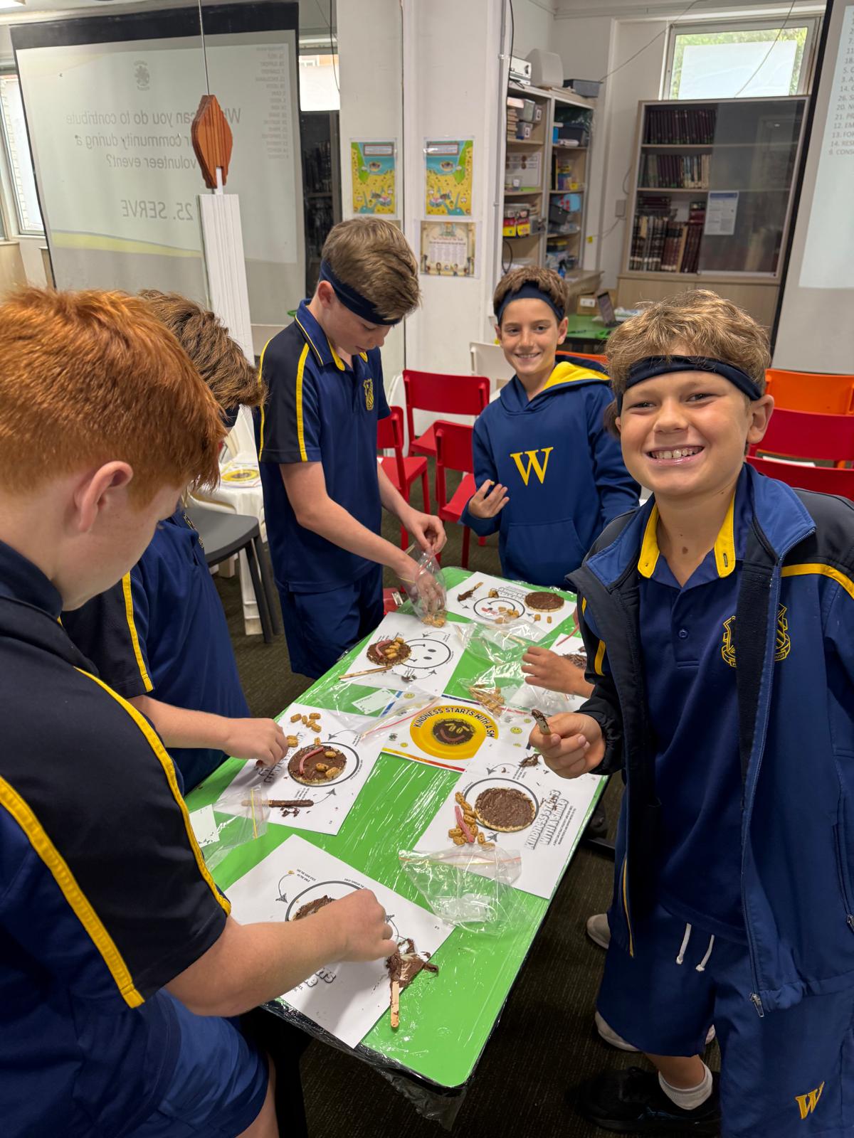 A group of boys in blue and yellow uniforms smile and work on craft projects at a green table in a classroom, with art supplies, snacks, and paper projects in front of them. Bookshelves and posters are in the background.