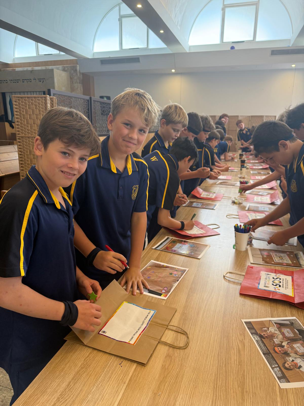 A group of smiling school children in navy uniforms stand around a long table, drawing and writing on paper bags in a bright classroom with large windows.